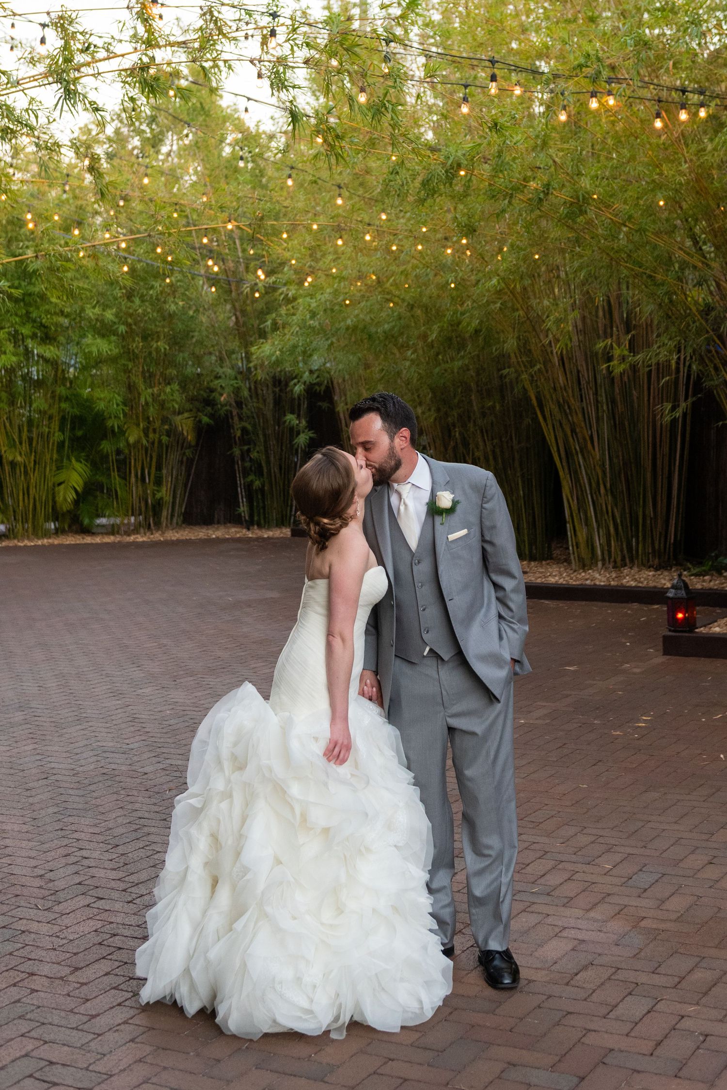 Newlyweds share a tender kiss on brick patio surrounded by bamboo and warm string lights at reception.