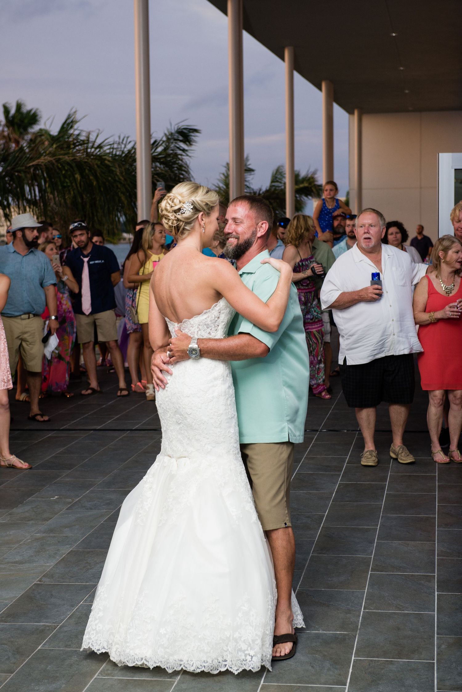 Bride and groom share first dance in outdoor venue, guests watching, bride in lace mermaid gown.