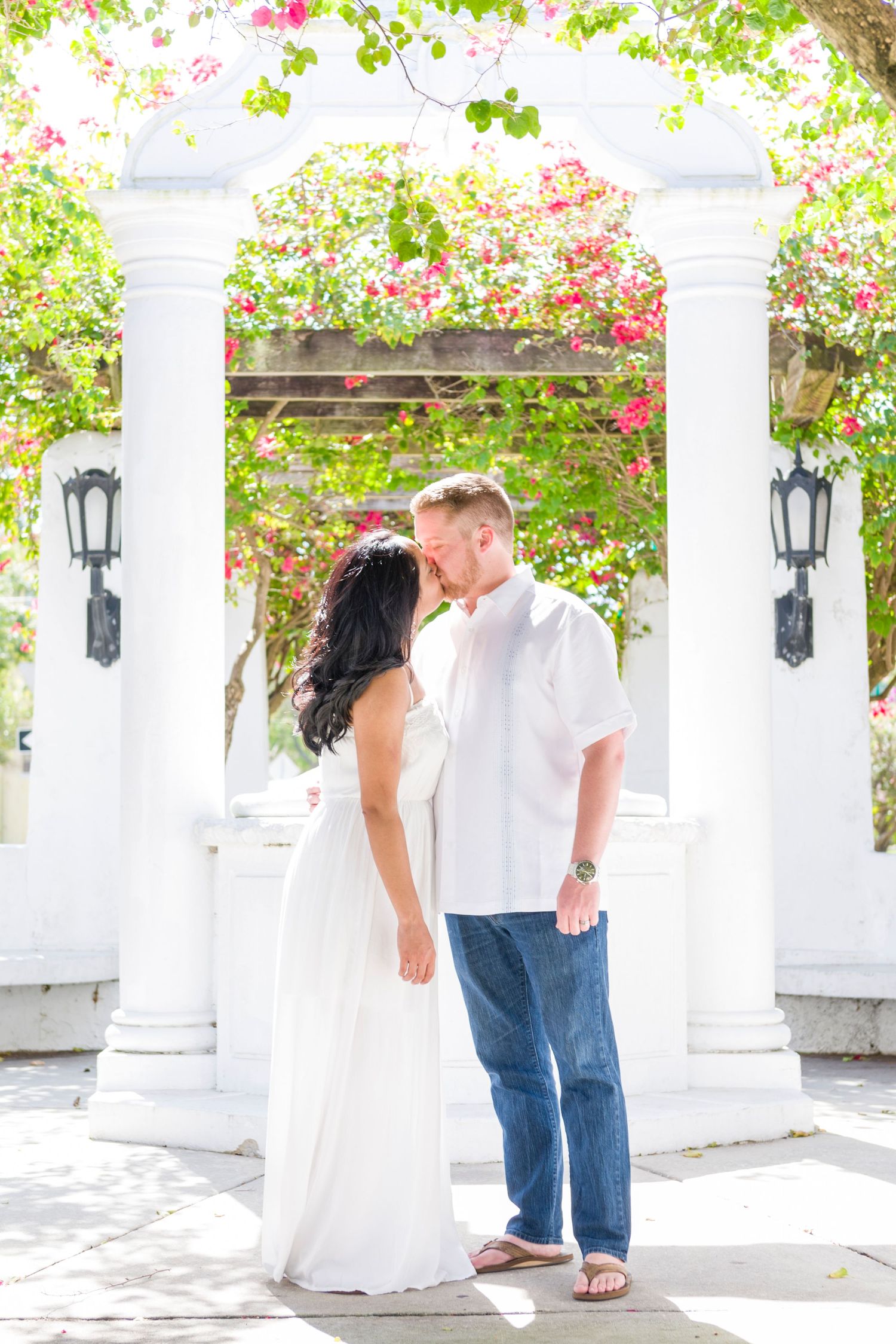 Couple sharing a romantic moment under a white pergola adorned with pink flowers, wearing white and jeans.