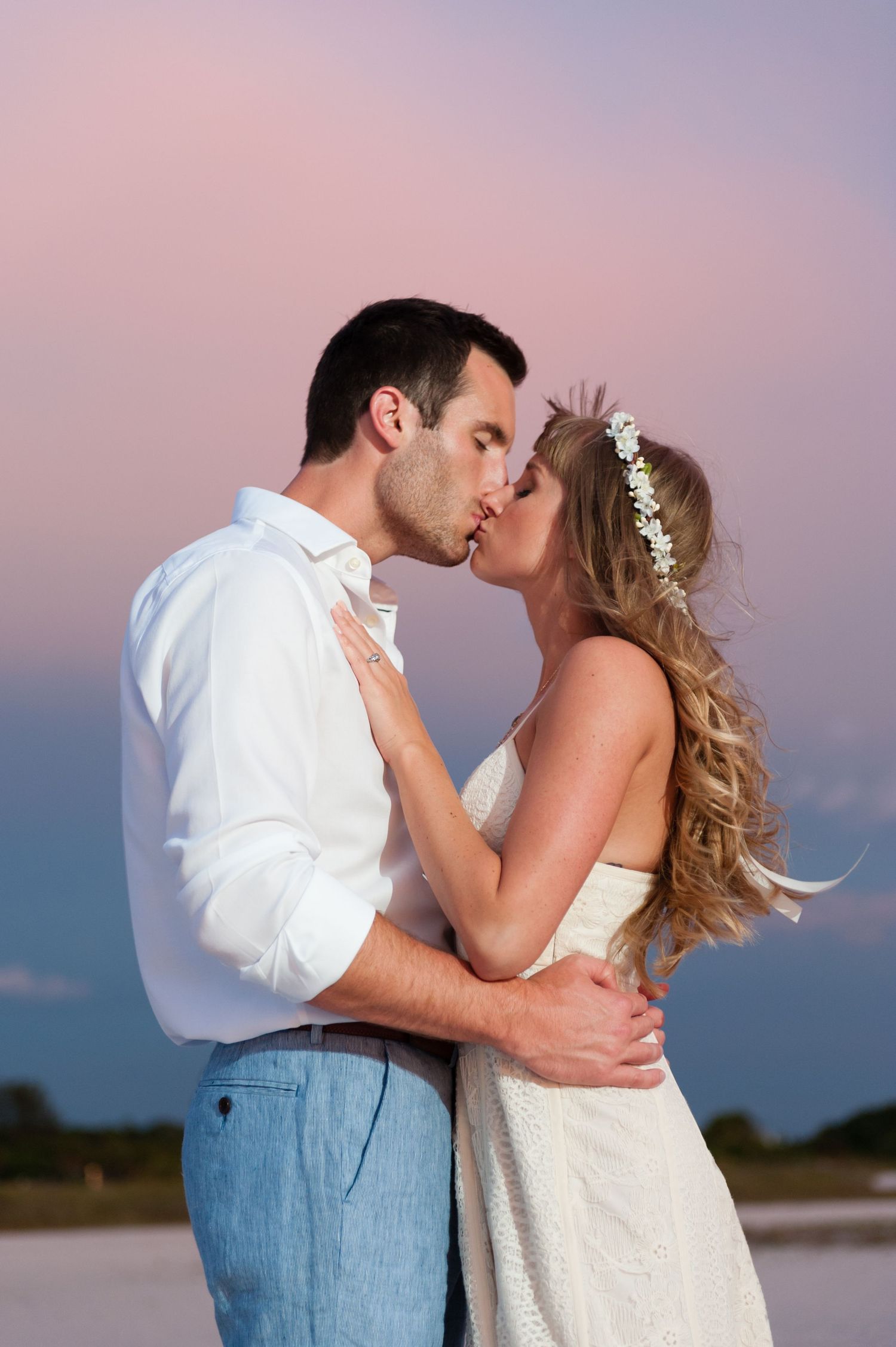 A bride and groom share a romantic kiss at sunset on a beach, she wearing a white dress and floral hair wreath.