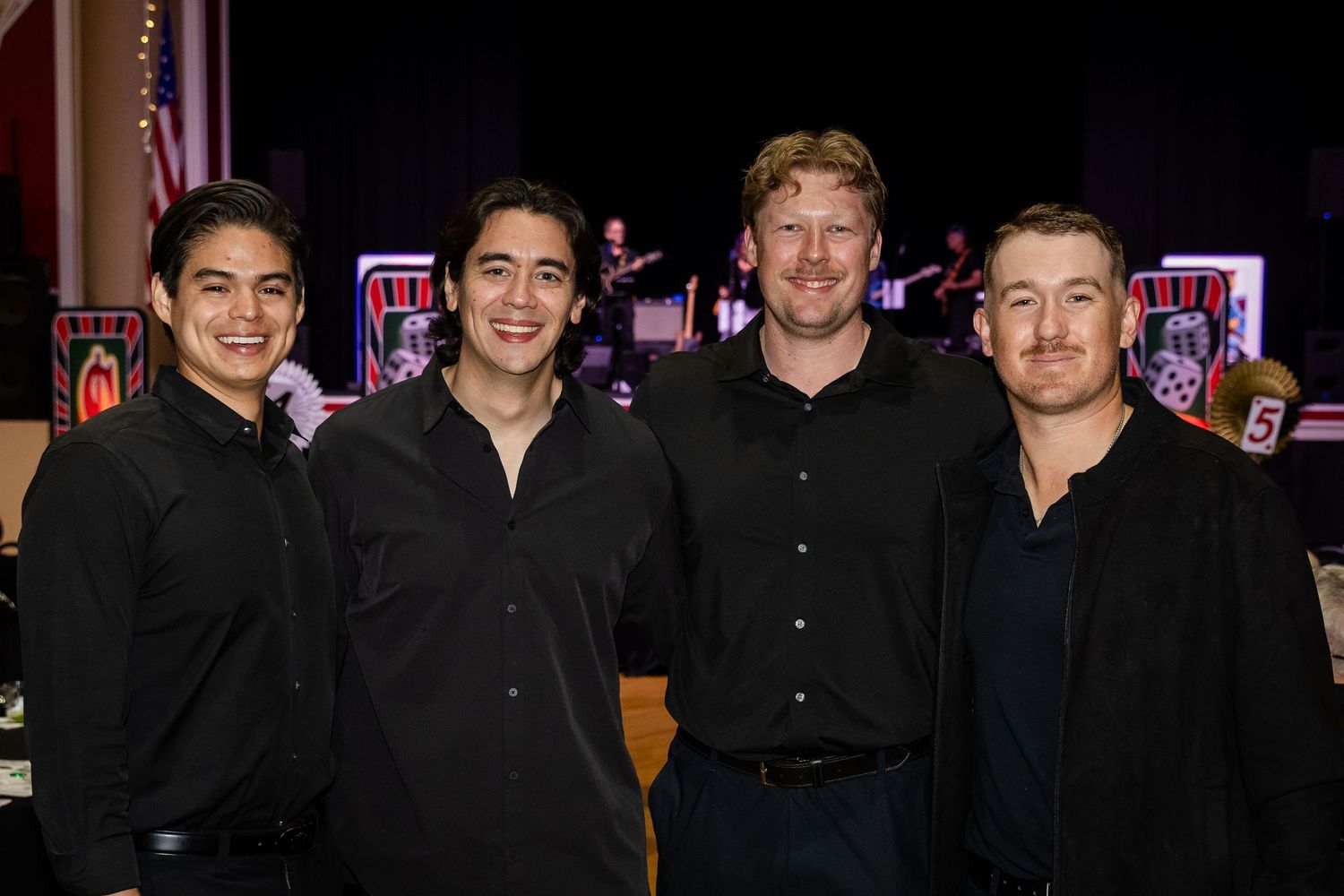 Four men in black shirts smiling together at a formal event with stage lighting in the background.