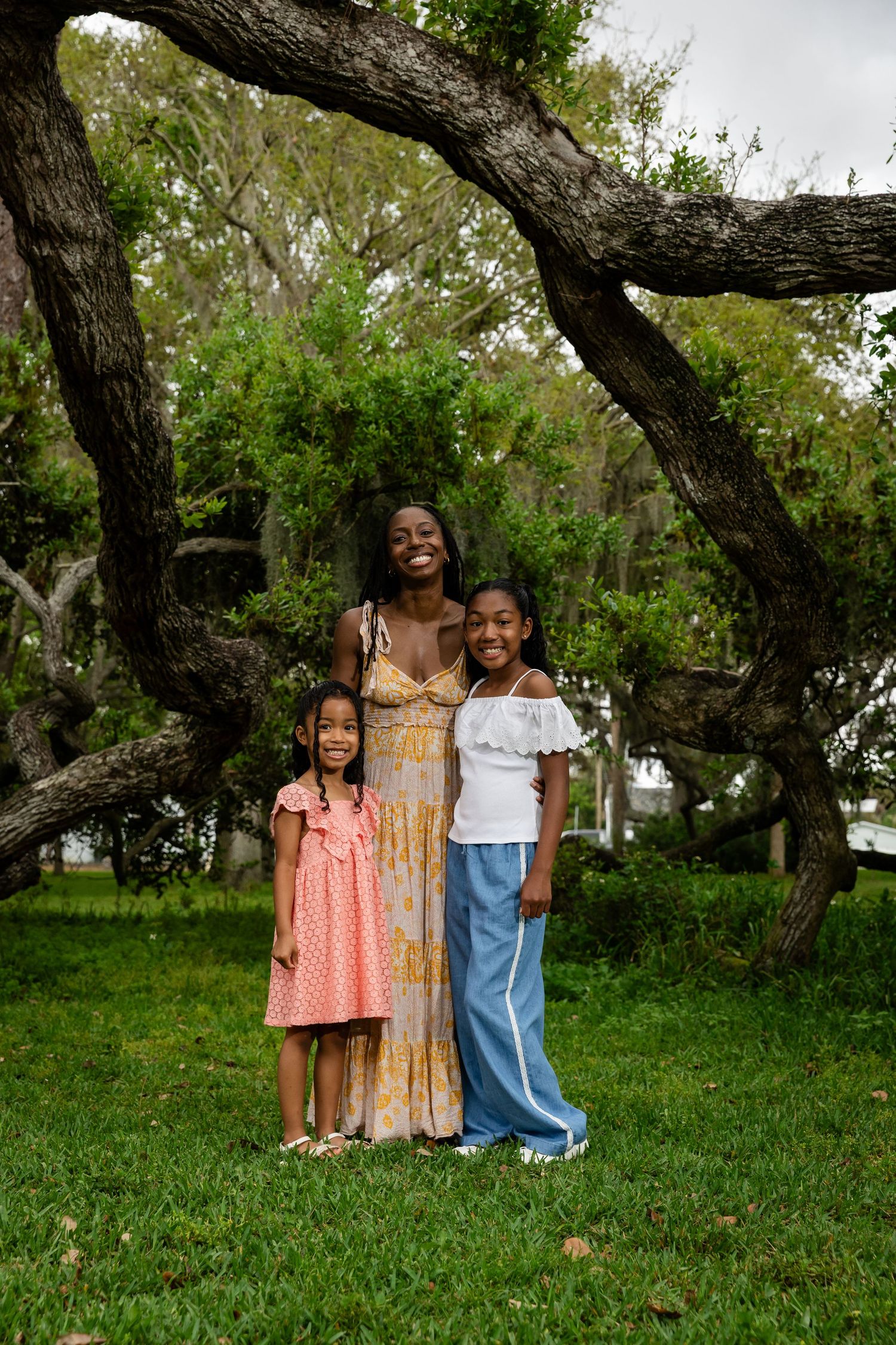 A mother and two daughters smile together outdoors under a large tree on green grass.