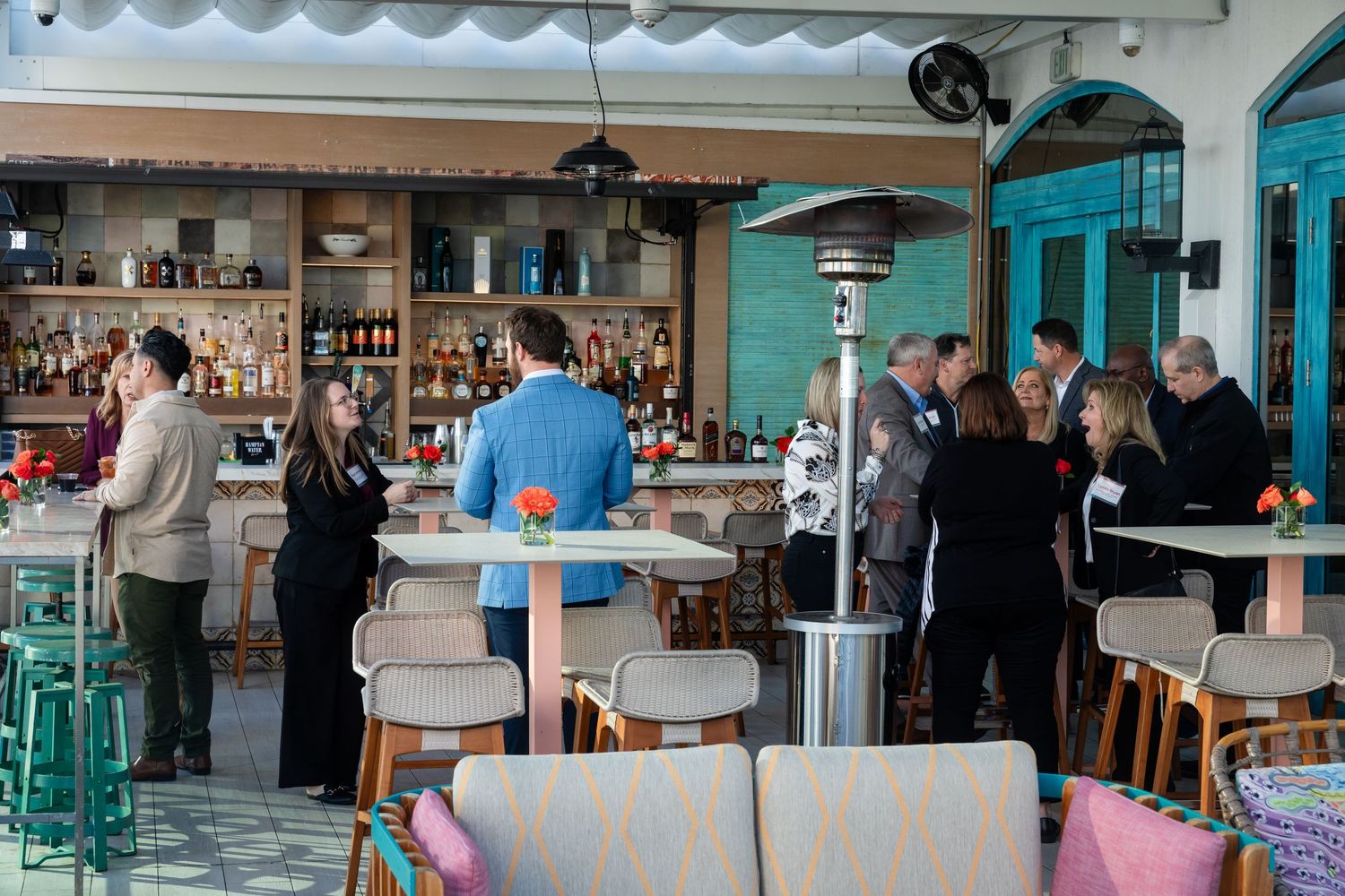 Guests mingle at an outdoor rooftop bar with seating areas and a patio heater during a social event.
