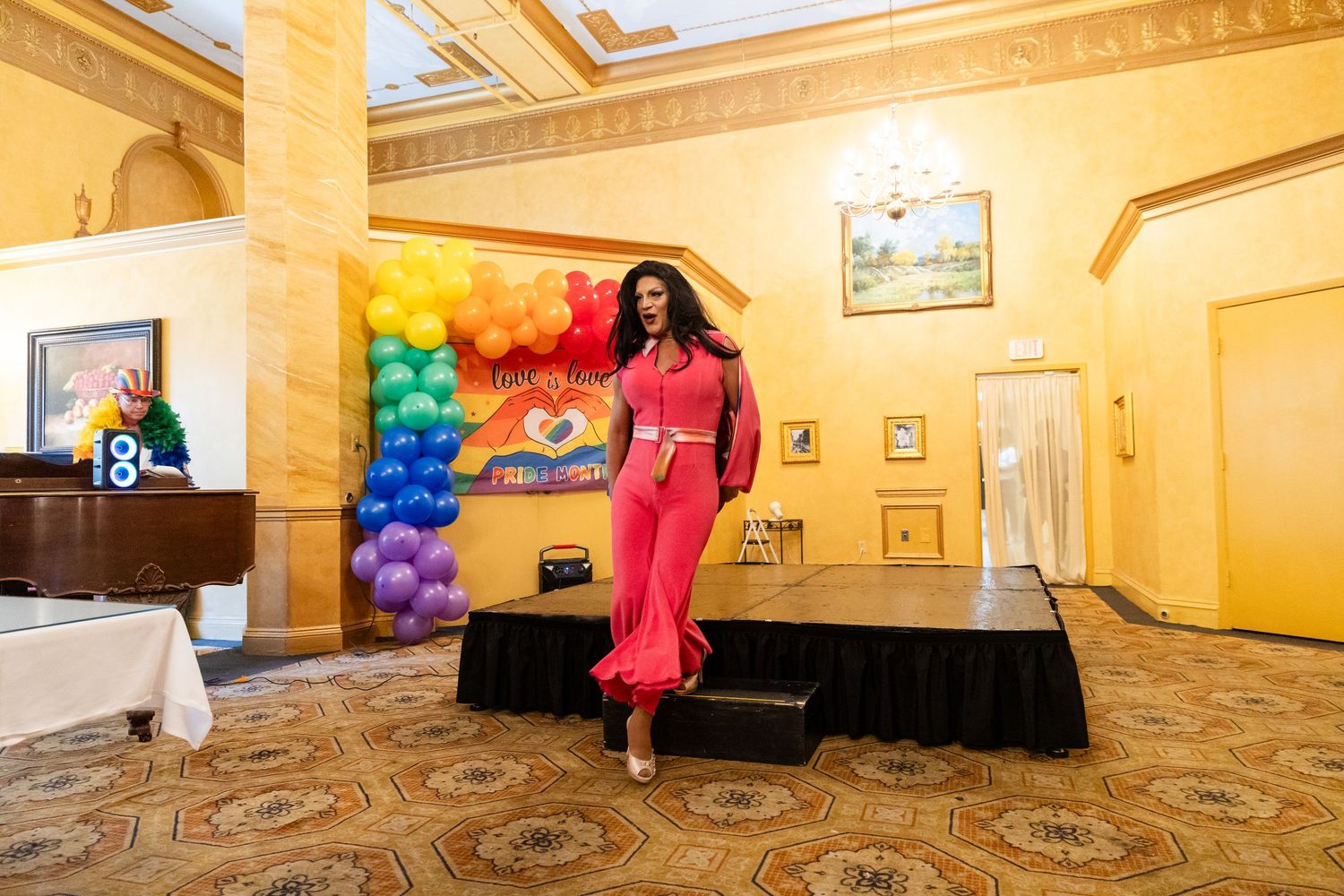 A model in a pink outfit walks the runway at a Pride-themed fashion show with rainbow balloon arch backdrop.