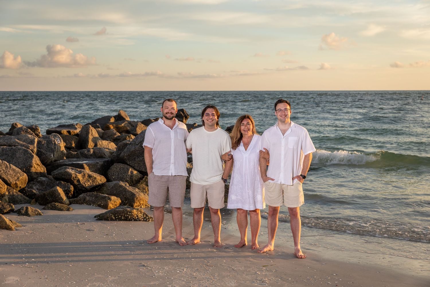 Happy family of four in white and khaki outfits posing together on sunny beach by jetty rocks.