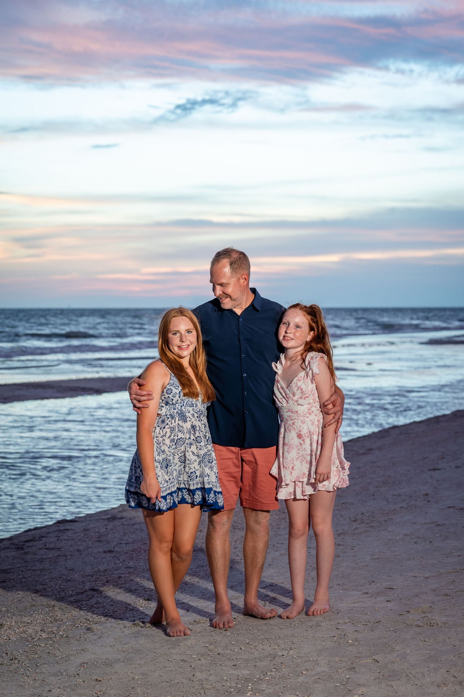Father and two daughters pose together on a sandy beach at sunset with colorful sky and ocean waves behind them.