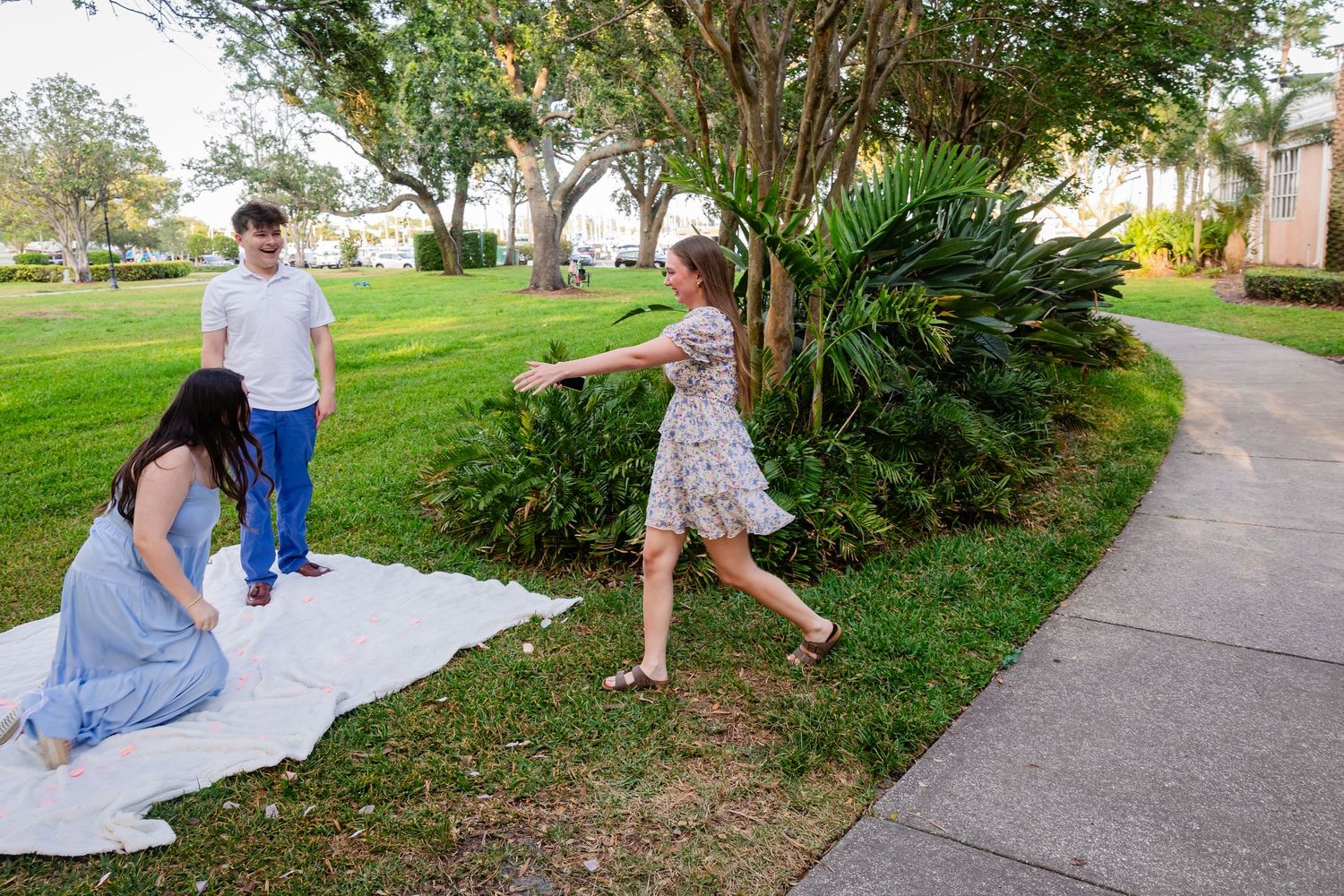 Three people setting up a white blanket on green grass near tropical shrubs and a sidewalk outdoors.