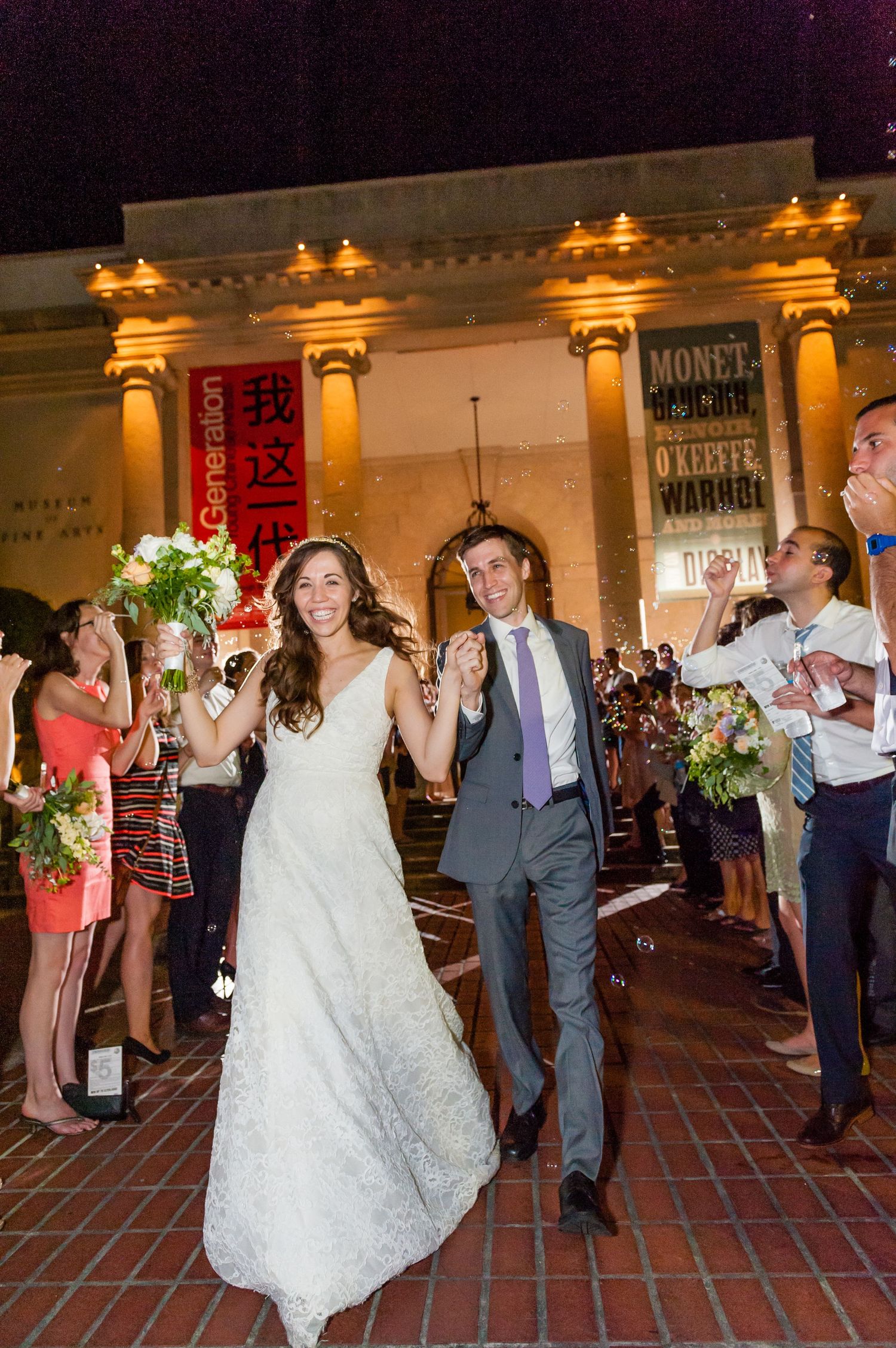 Joyful newlyweds exit wedding venue as guests celebrate outside illuminated museum at night.