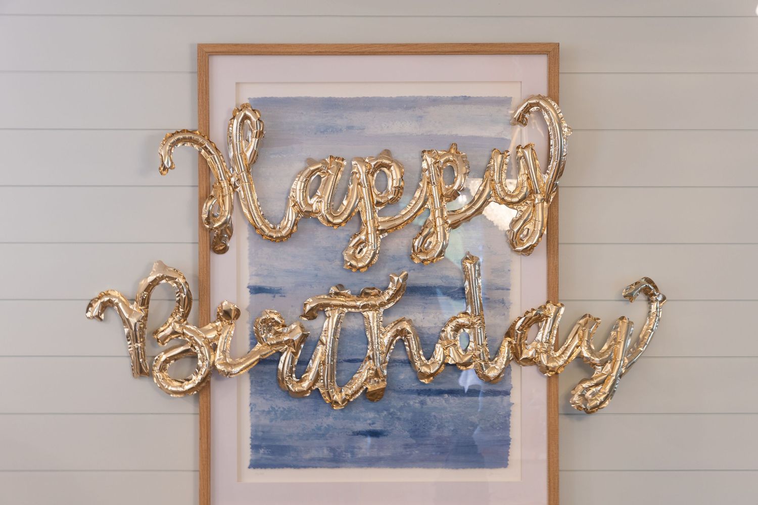Gold foil Happy Birthday balloon letters displayed against a watercolor painting on a white shiplap wall.