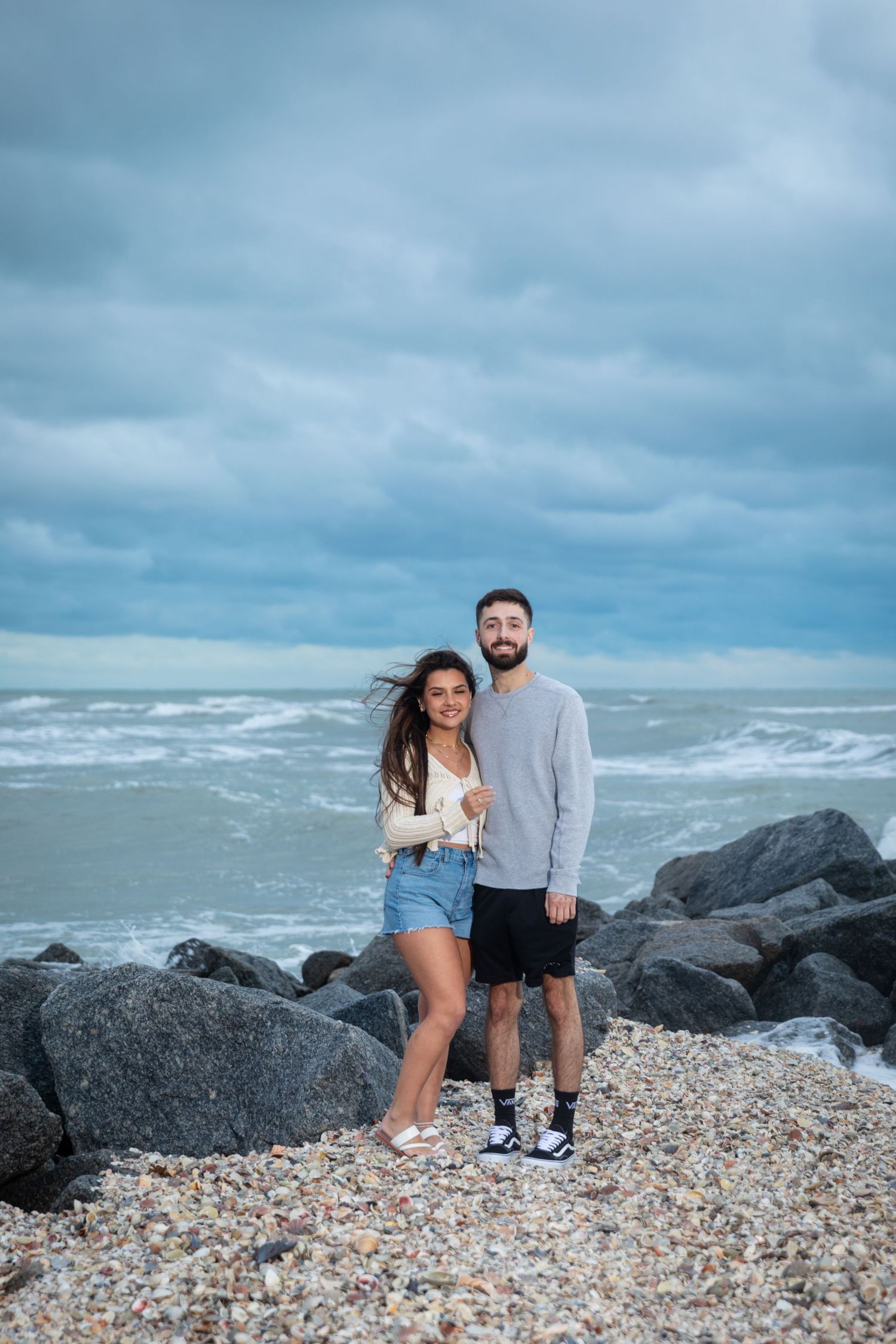 Happy couple posing together on a rocky shoreline with dramatic cloudy skies and ocean waves.