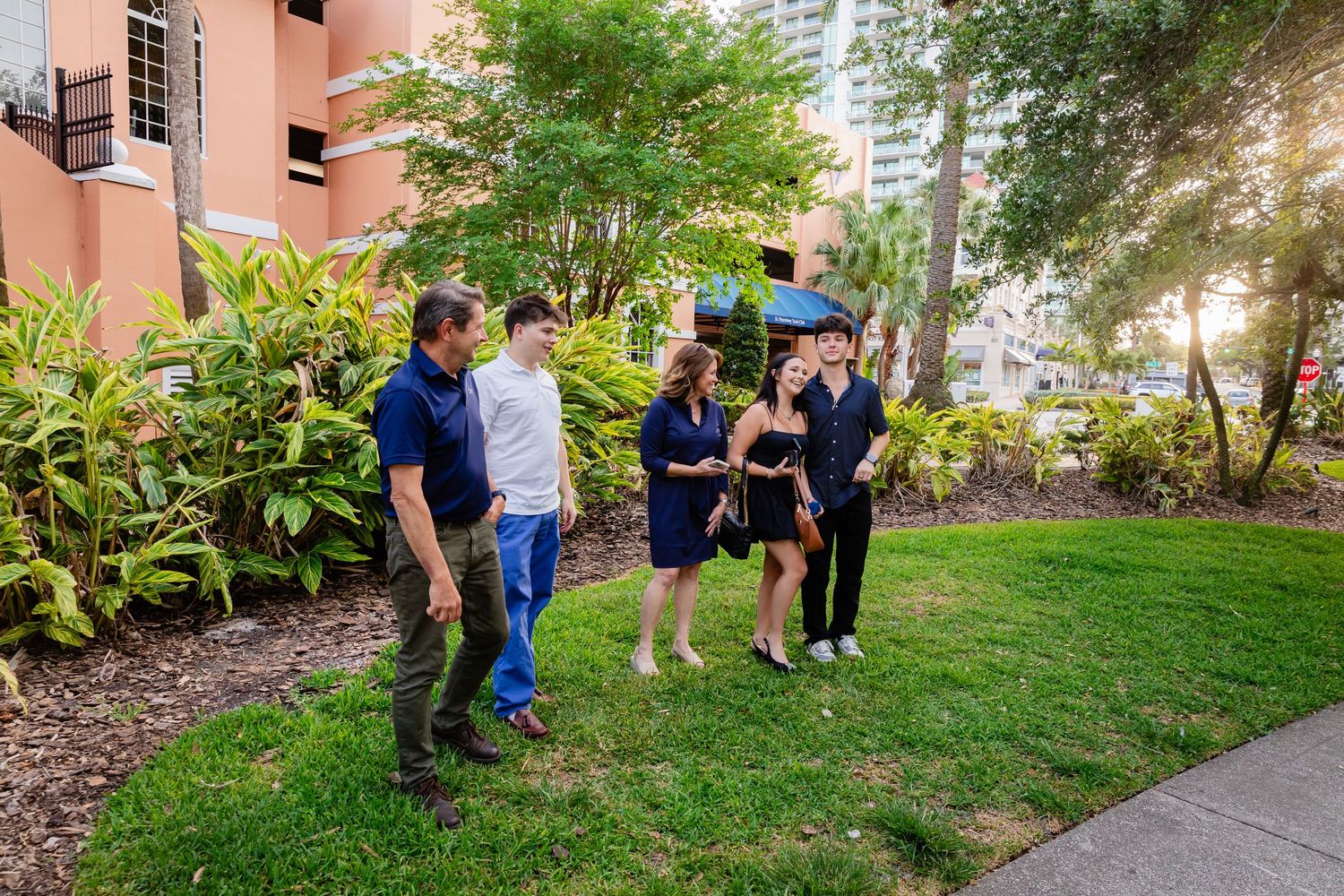 A family group of five people conversing on a green lawn surrounded by tropical trees and urban buildings at sunset.
