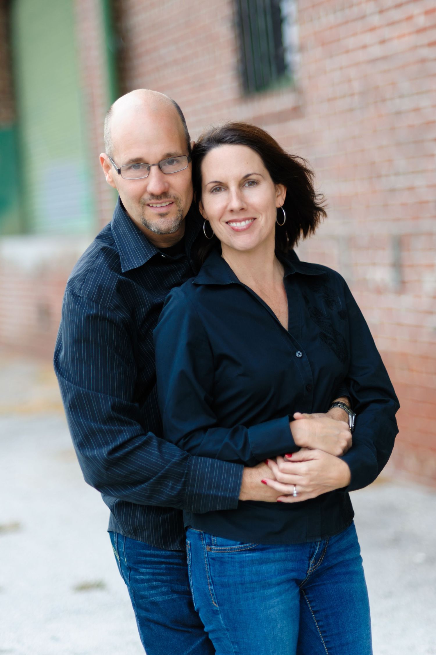 A smiling couple in black shirts embracing outdoors near a brick wall, posing for a portrait photo.