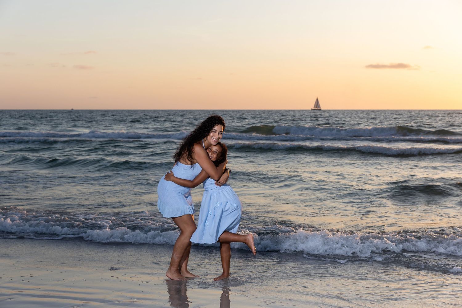 Two women in blue dresses embrace joyfully on a beach at sunset with a sailboat in the distance.