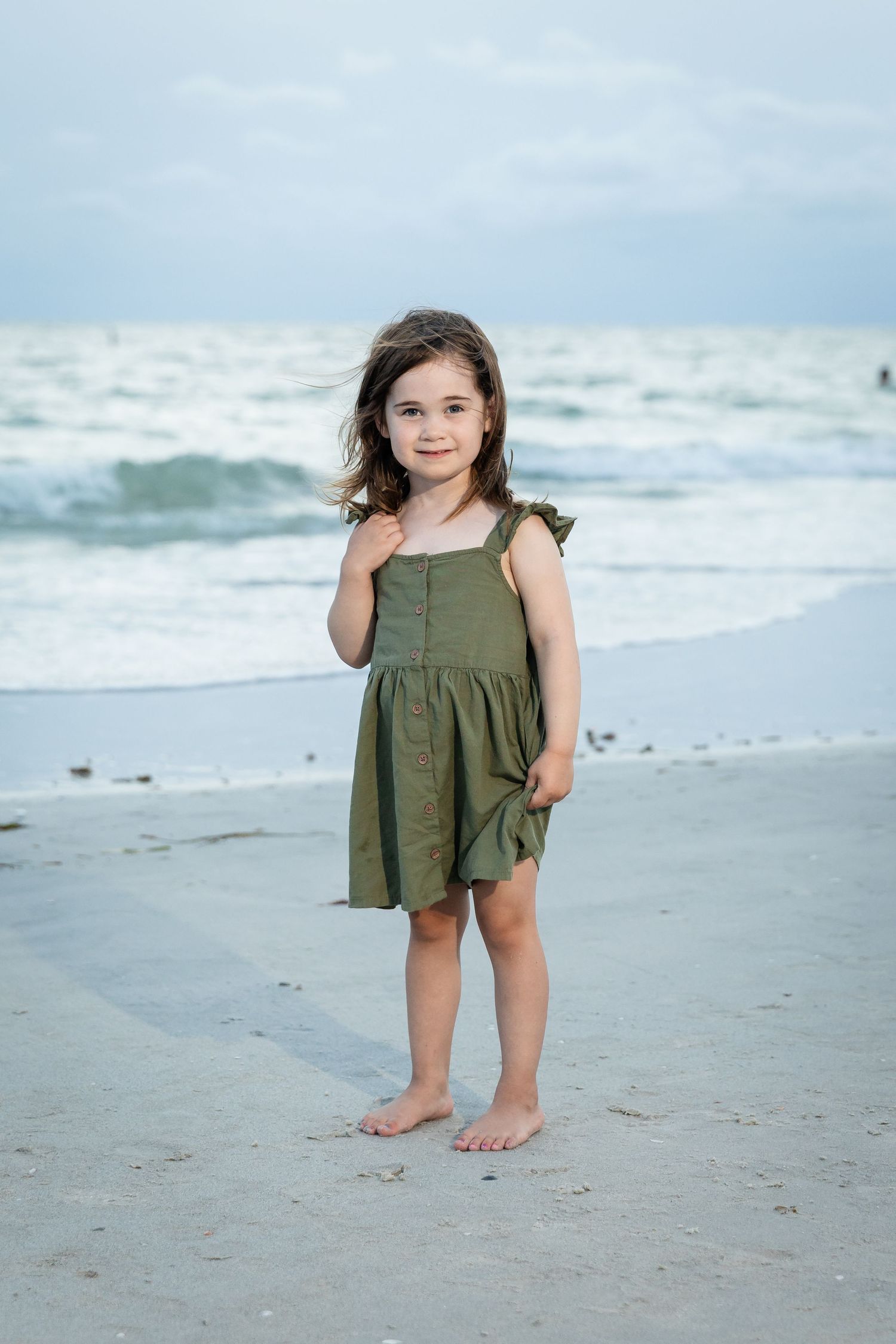 Young girl in olive green dress standing barefoot on sandy beach with ocean waves in background.