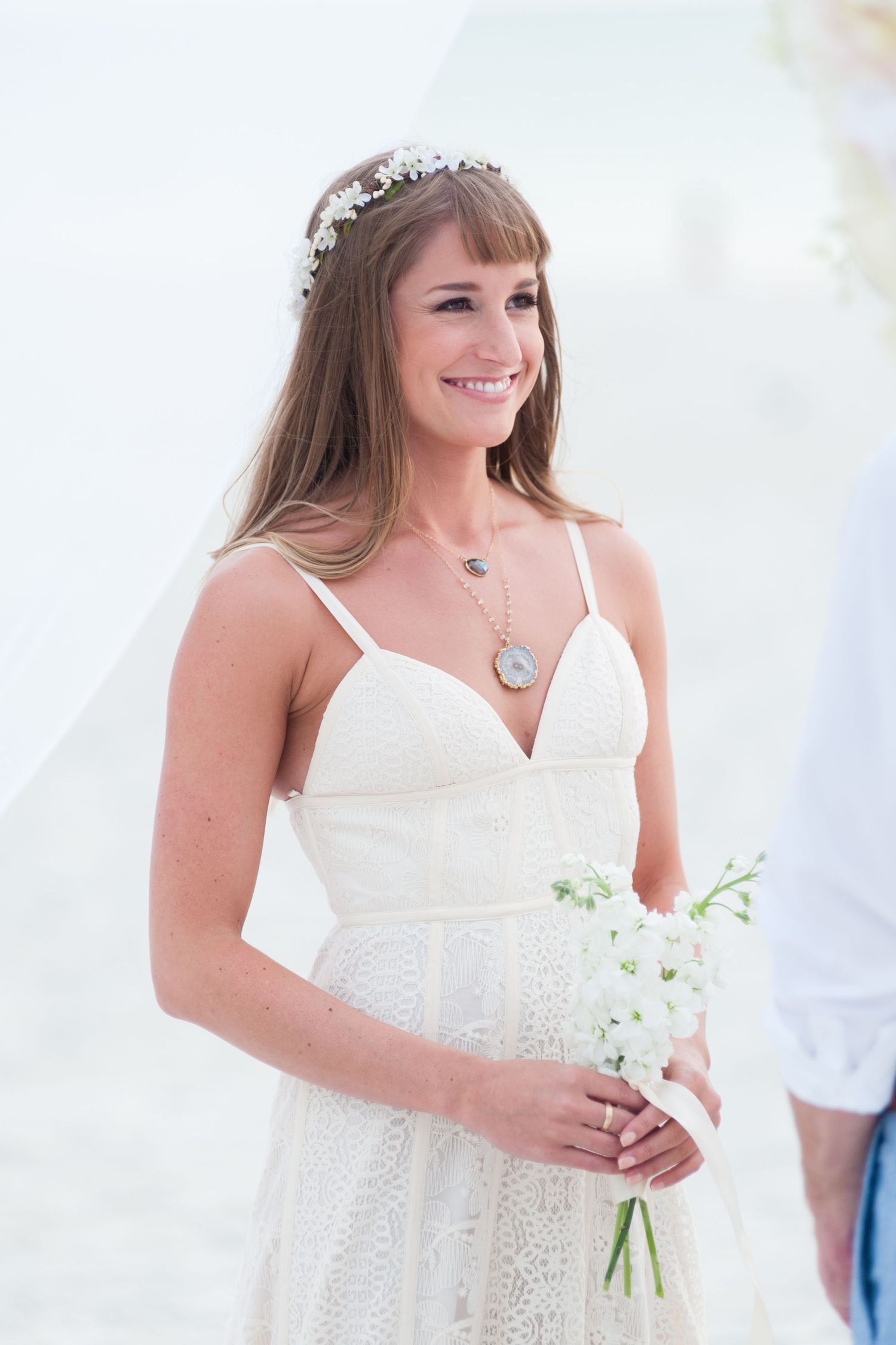 Smiling bride wearing a white lace dress and floral crown holding a white bouquet at beach wedding.