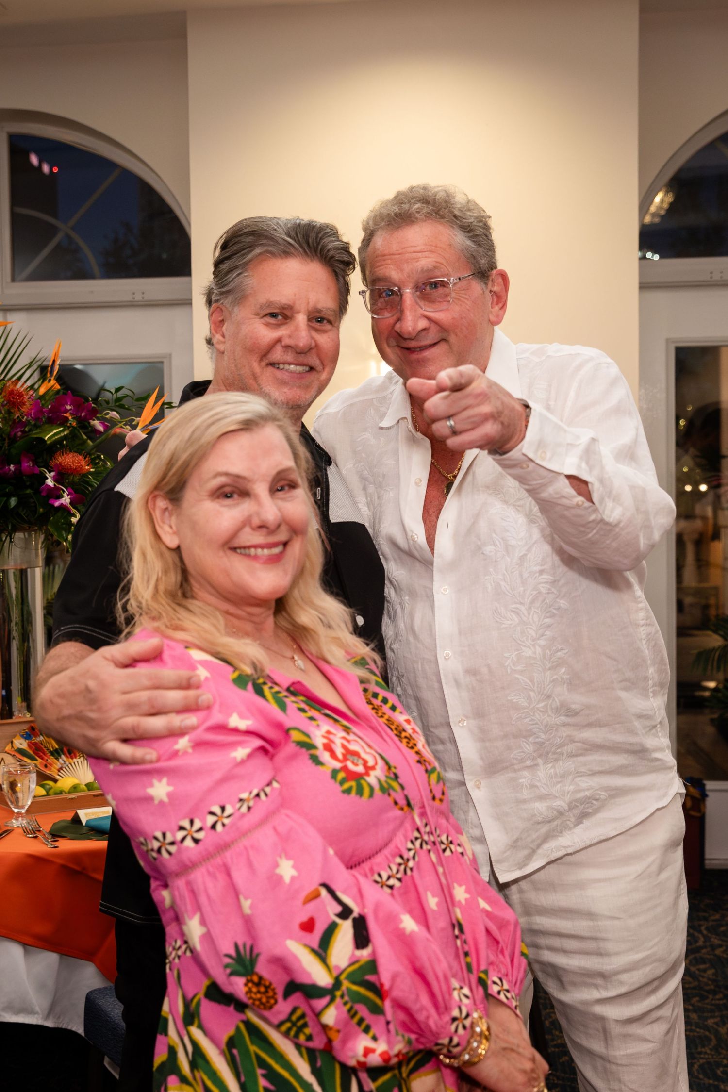 Three smiling adults pose together at a festive indoor event, with a woman in pink floral dress flanked by two men.
