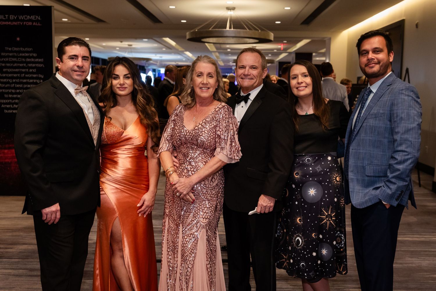 Six elegantly dressed guests posing together at a formal gala event, wearing evening gowns and suits.