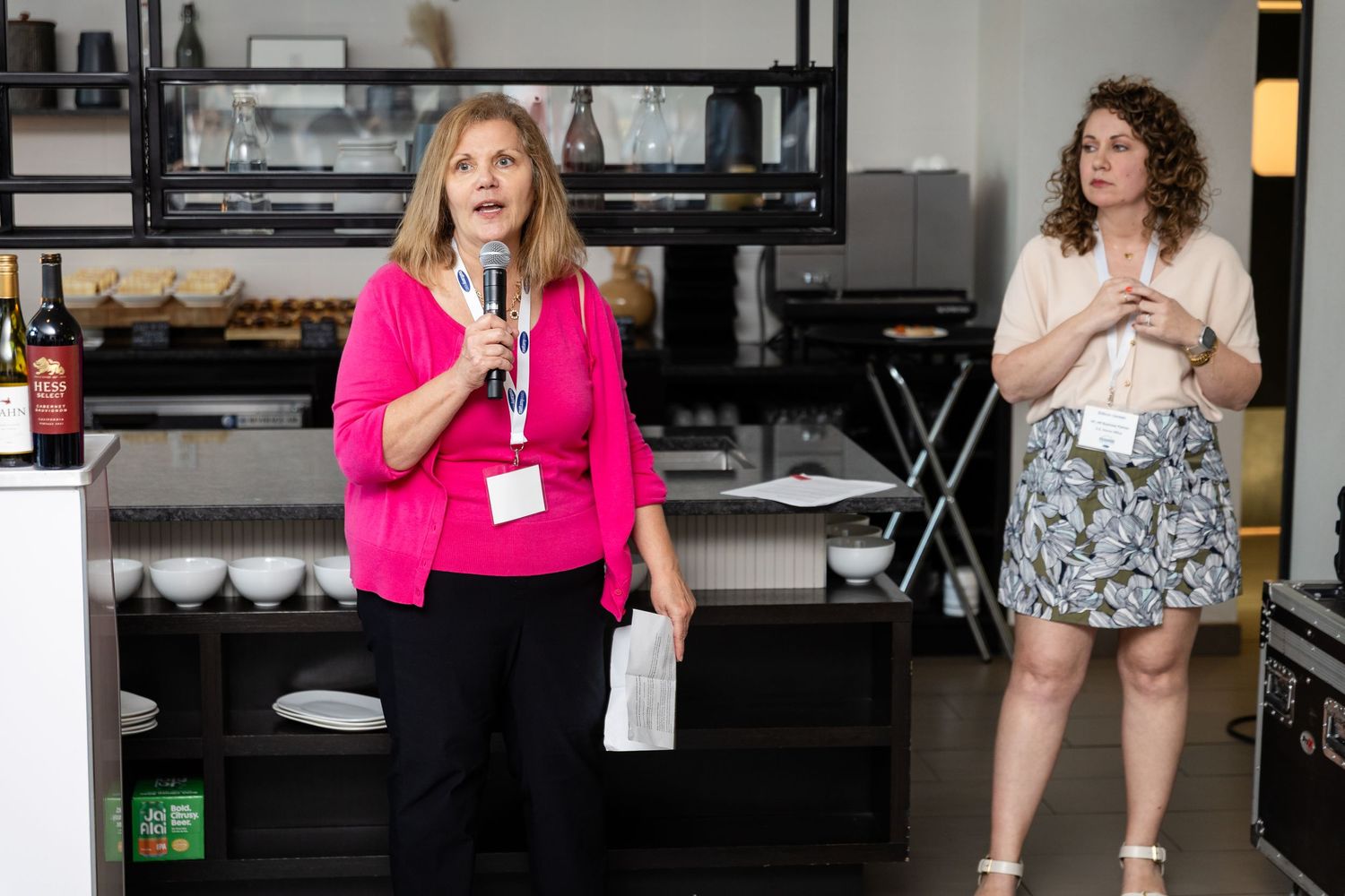 Woman in pink blouse speaks at indoor event while colleague in floral skirt stands nearby at catered venue.