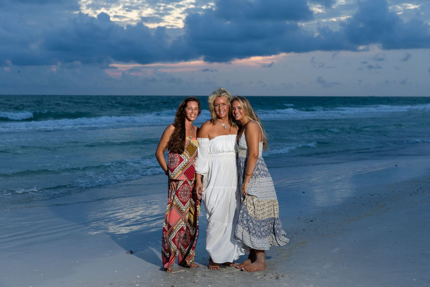 Three smiling women in flowing dresses pose together on a white sand beach under dramatic clouds.