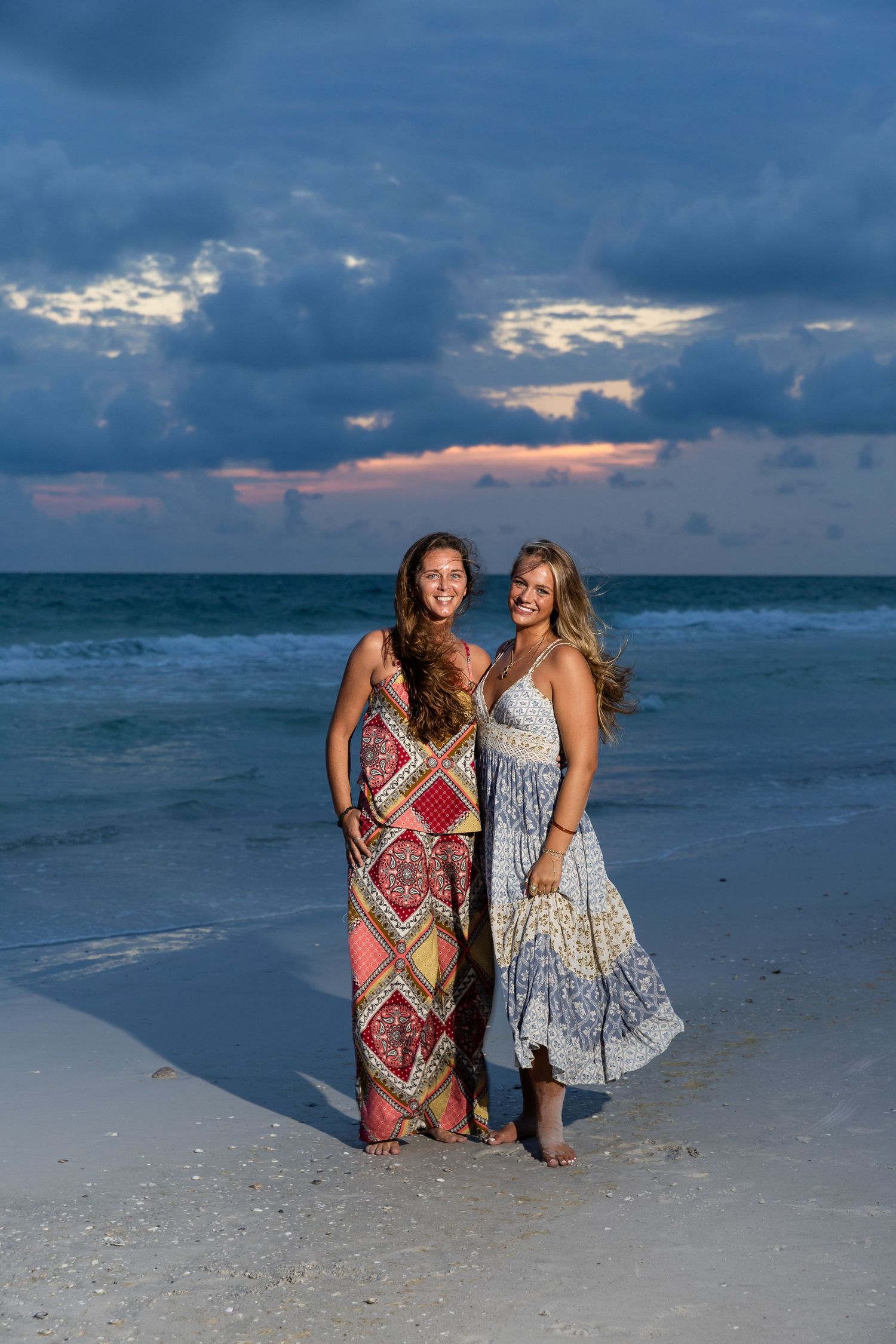 Two women in boho maxi dresses smile on a sandy beach at sunset with dramatic cloudy skies and ocean waves.