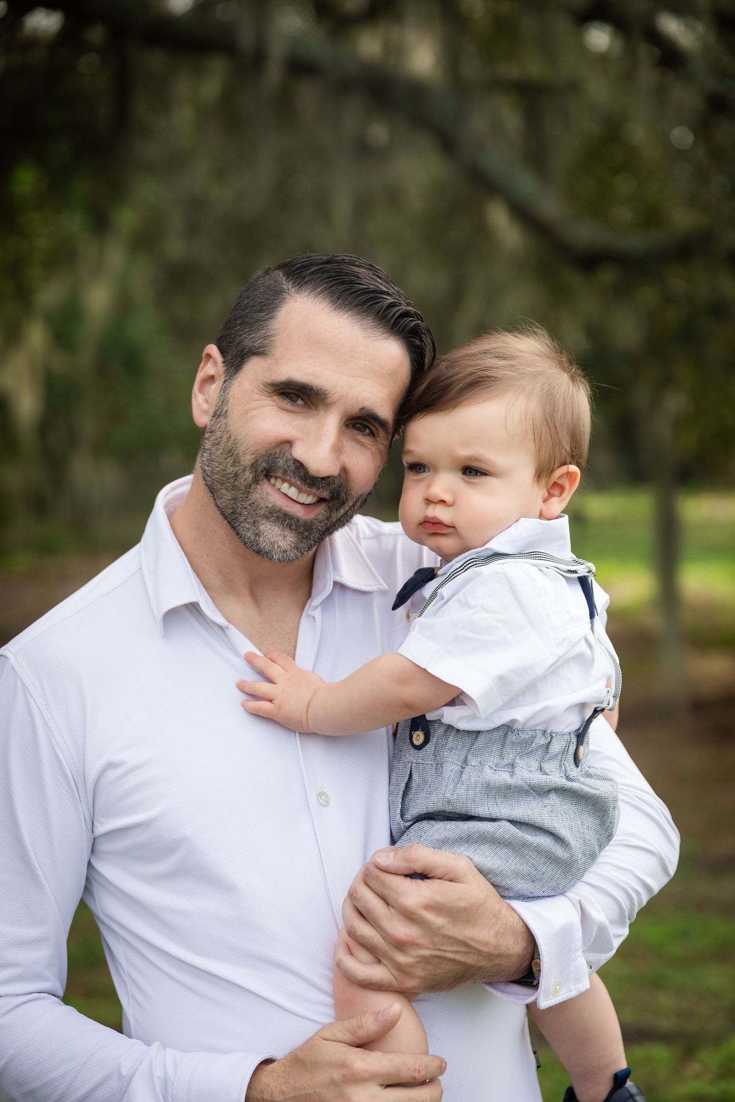 A smiling bearded man in a white shirt holds a baby boy in suspenders outdoors among moss-covered trees.