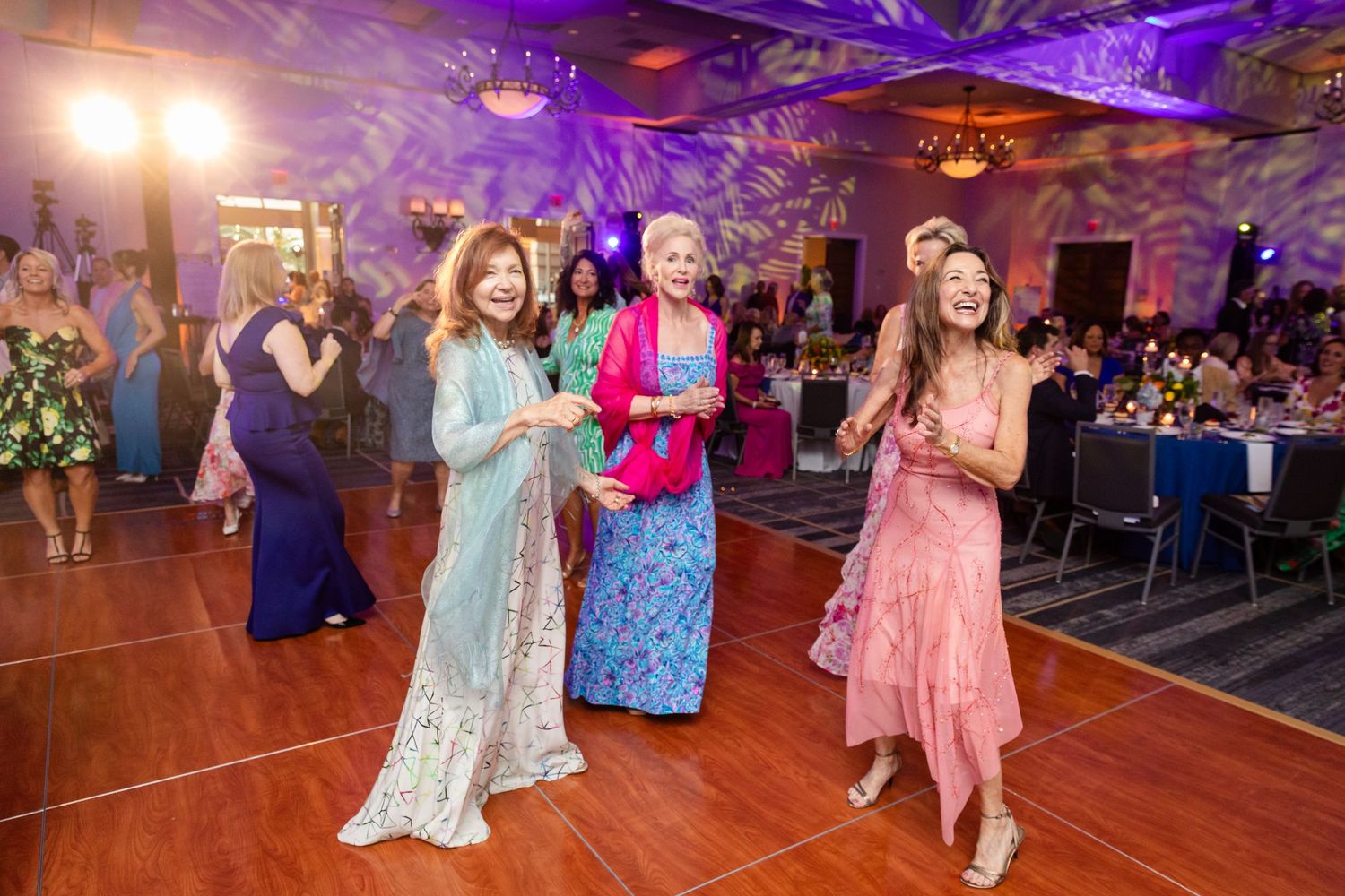 Three women dancing joyfully in elegant dresses at a wedding reception with purple lighting.