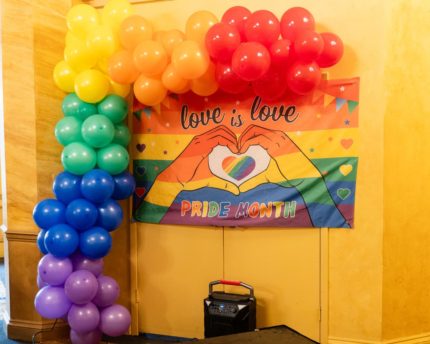 Rainbow balloon arch beside a colorful Pride Month banner reading love is love at an indoor celebration.