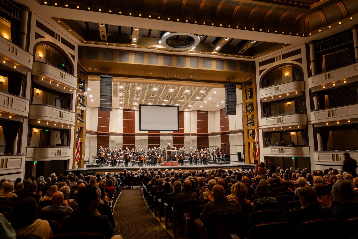 A packed concert hall with orchestra performing on stage, featuring elegant balconies and warm lighting.