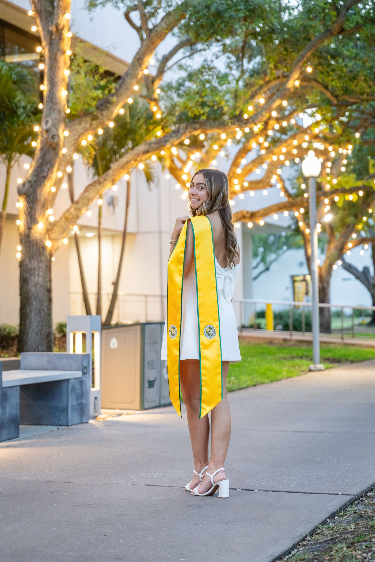 Young woman in white dress holding yellow graduation stole, posing on campus walkway with string lights in trees.