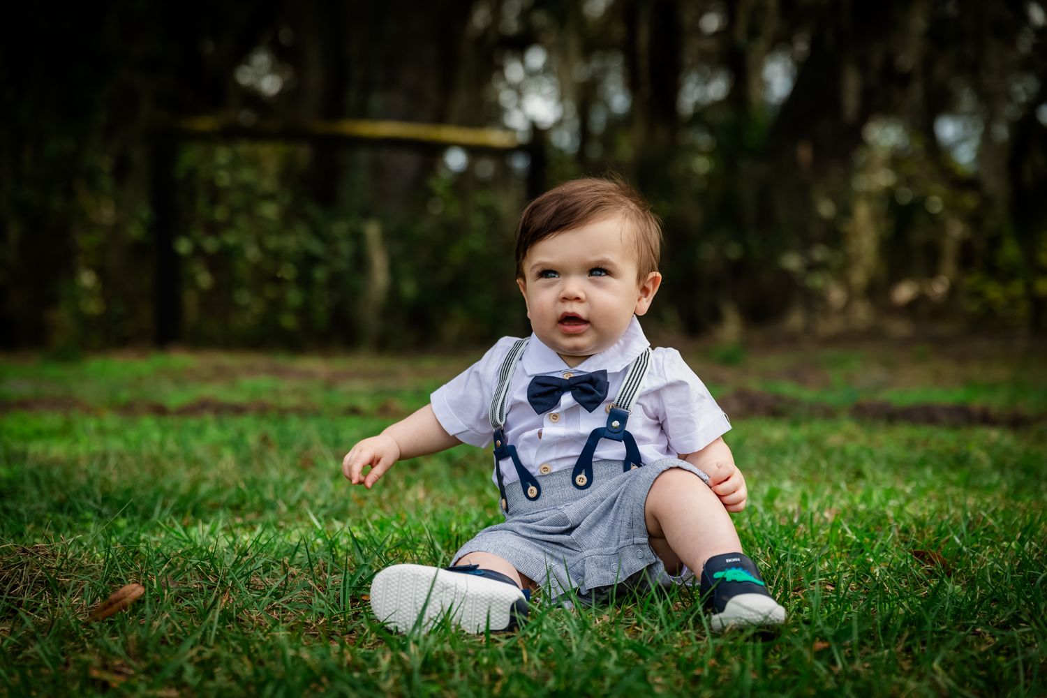 Adorable baby boy in suspenders shirt sitting on green grass outdoors, smiling in a park setting.