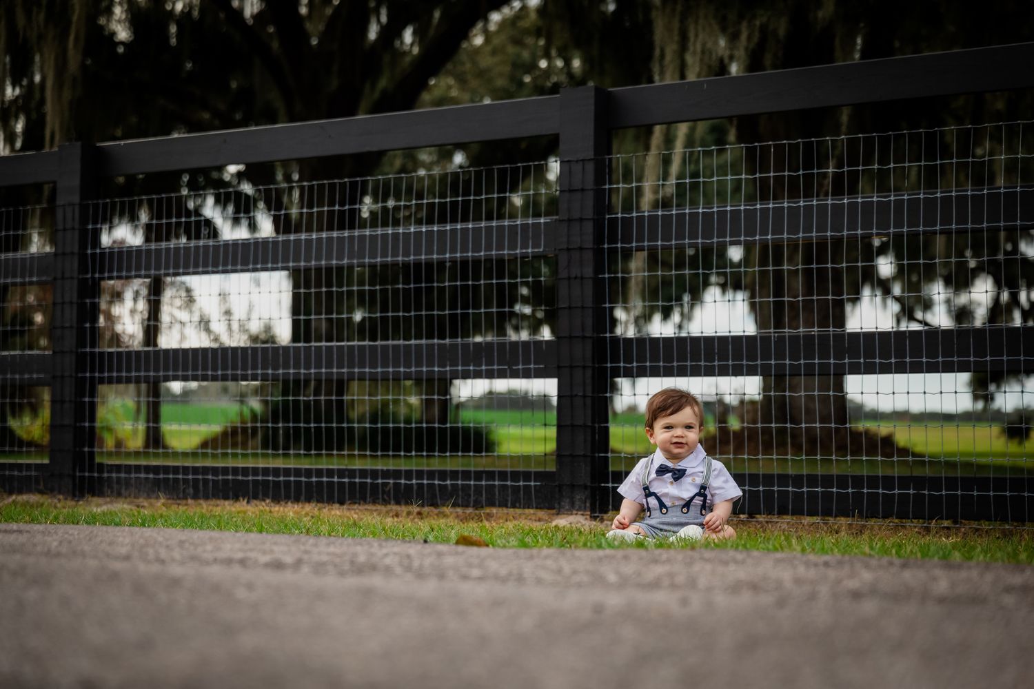 An adorable baby boy in suspenders sits alone on a path in front of a black metal farm fence.