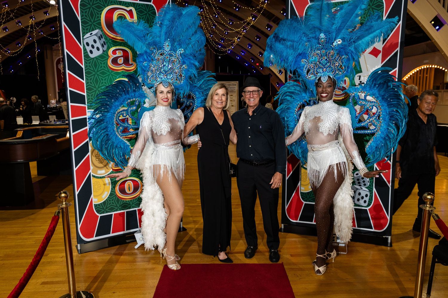 Two showgirls in blue feathered costumes pose with a couple on a red carpet at a casino-themed event.
