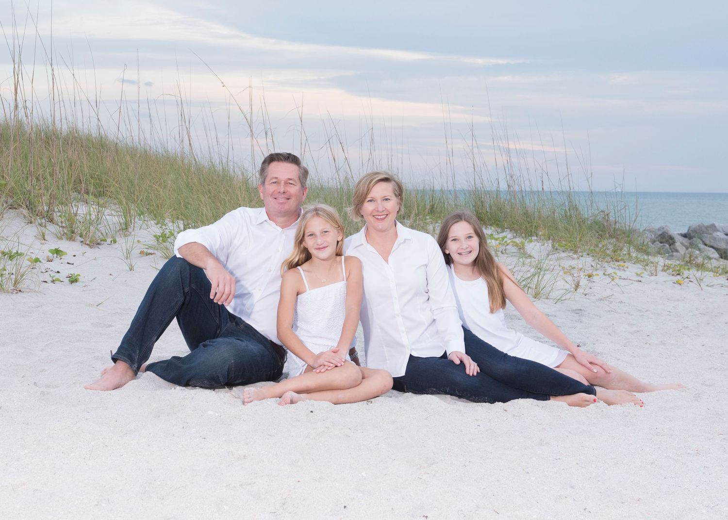 Family of four smiling together on a sandy beach, wearing white tops and navy bottoms, with sea grass and ocean in background.