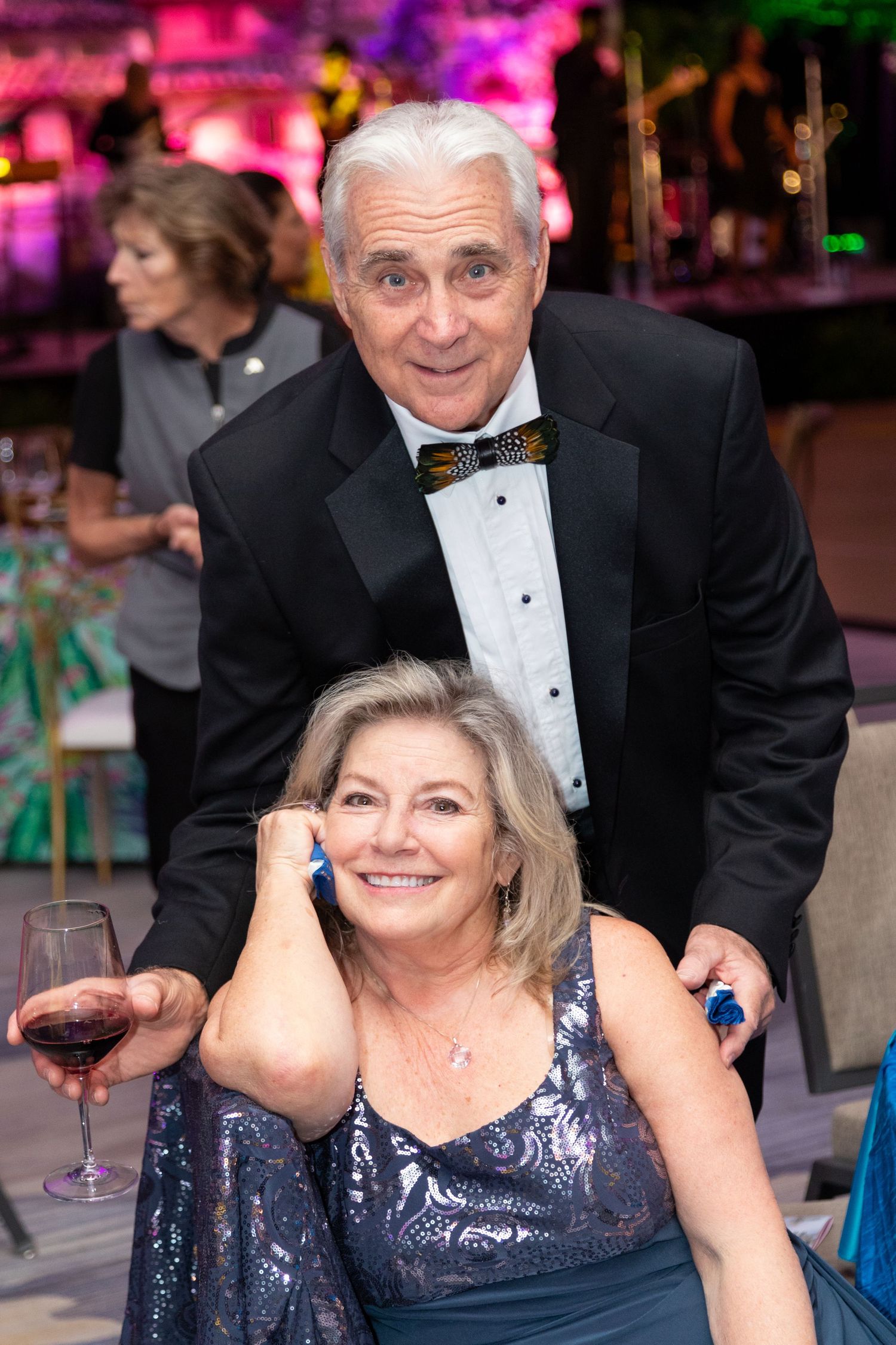 Elegant couple at formal gala, man in tuxedo with bow tie, woman in blue sequin dress holding red wine glass.
