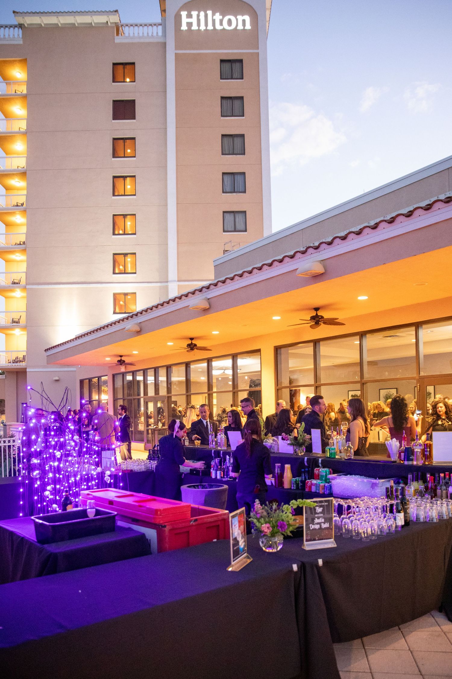 Outdoor evening reception at Hilton hotel featuring a bar setup with purple lighting and decorative trees.