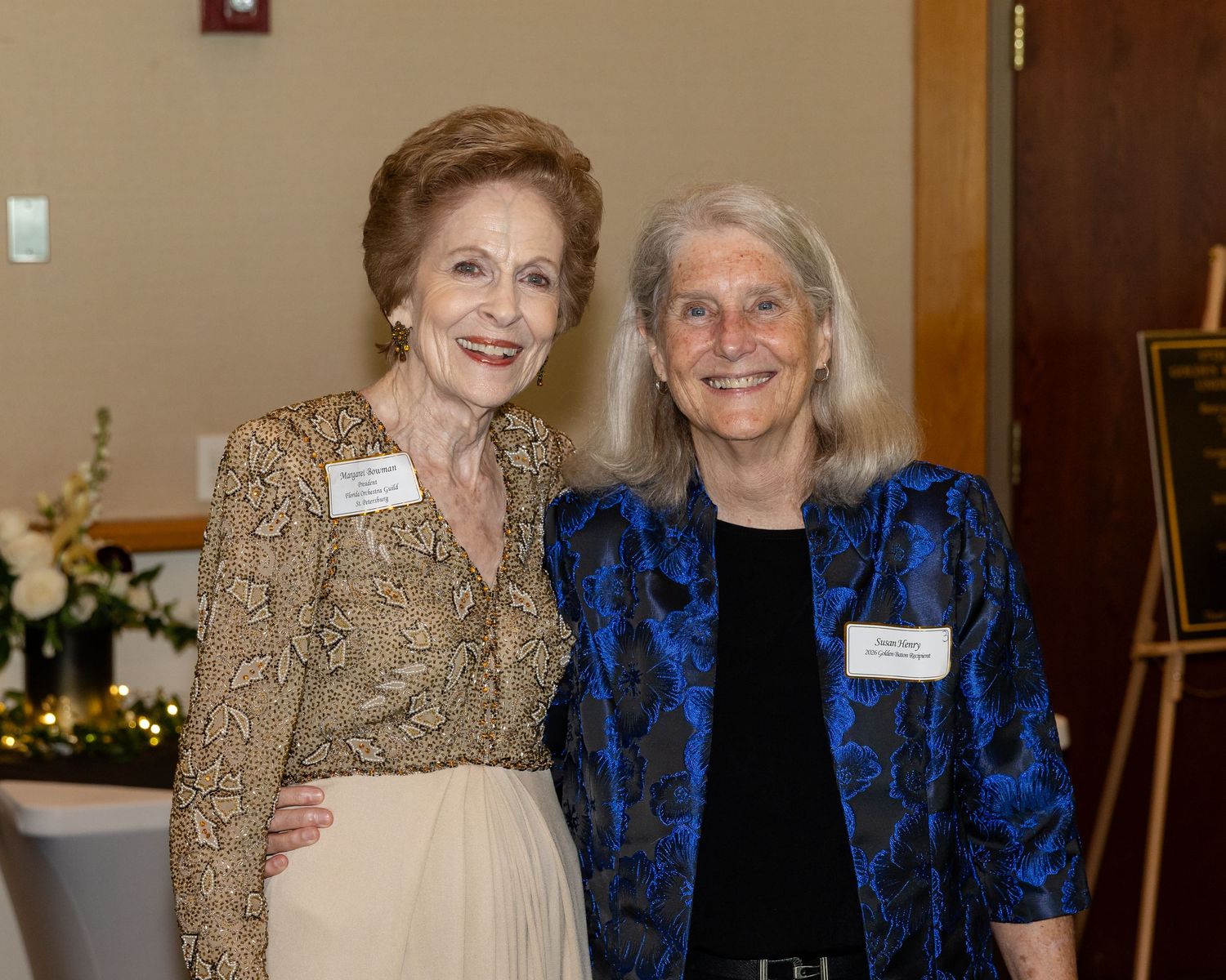Two elegantly dressed older women smiling together at a formal event, wearing name tags and festive attire.