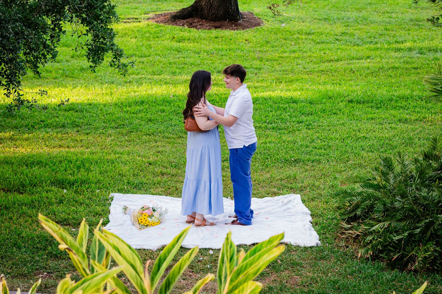 Couple standing together on a white blanket in a lush green park, sharing a romantic outdoor picnic moment.