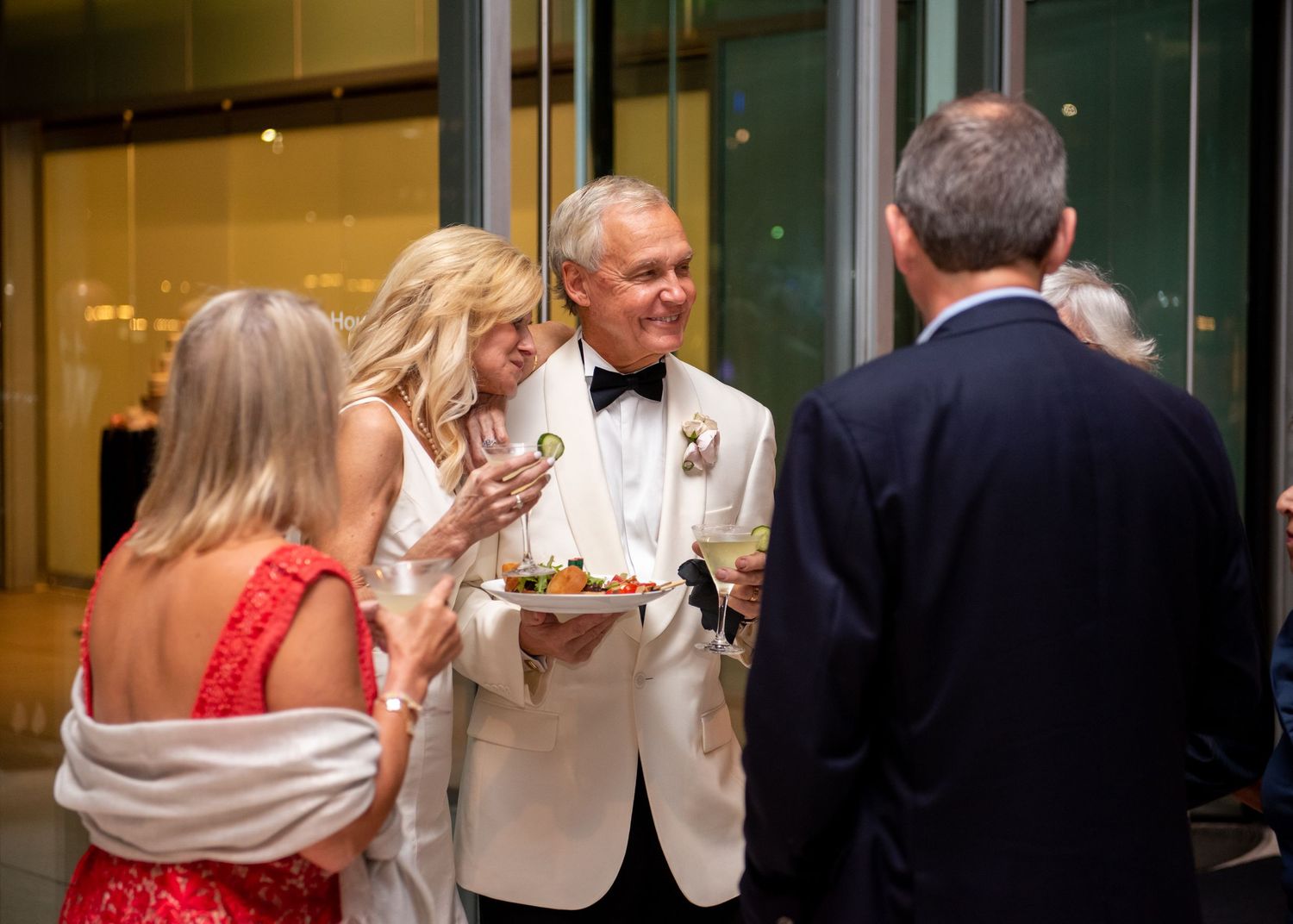Elegantly dressed guests socializing at a formal evening event, man in white tuxedo holding appetizer platter.