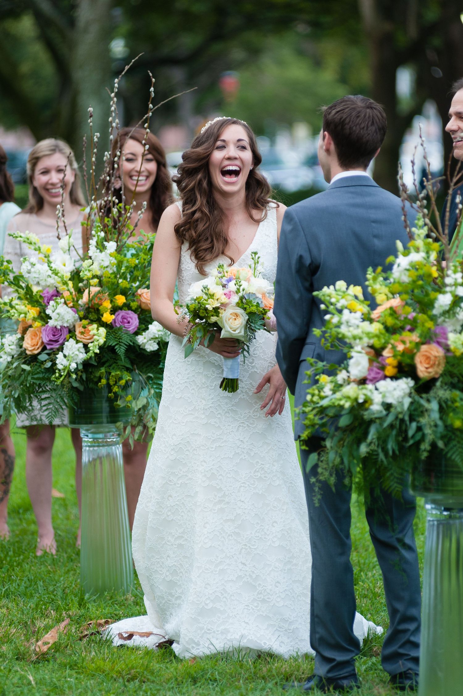 Joyful bride laughing at altar in white lace gown flanked by vibrant floral arrangements at outdoor wedding Museum of Fine Arts, St. Peters.