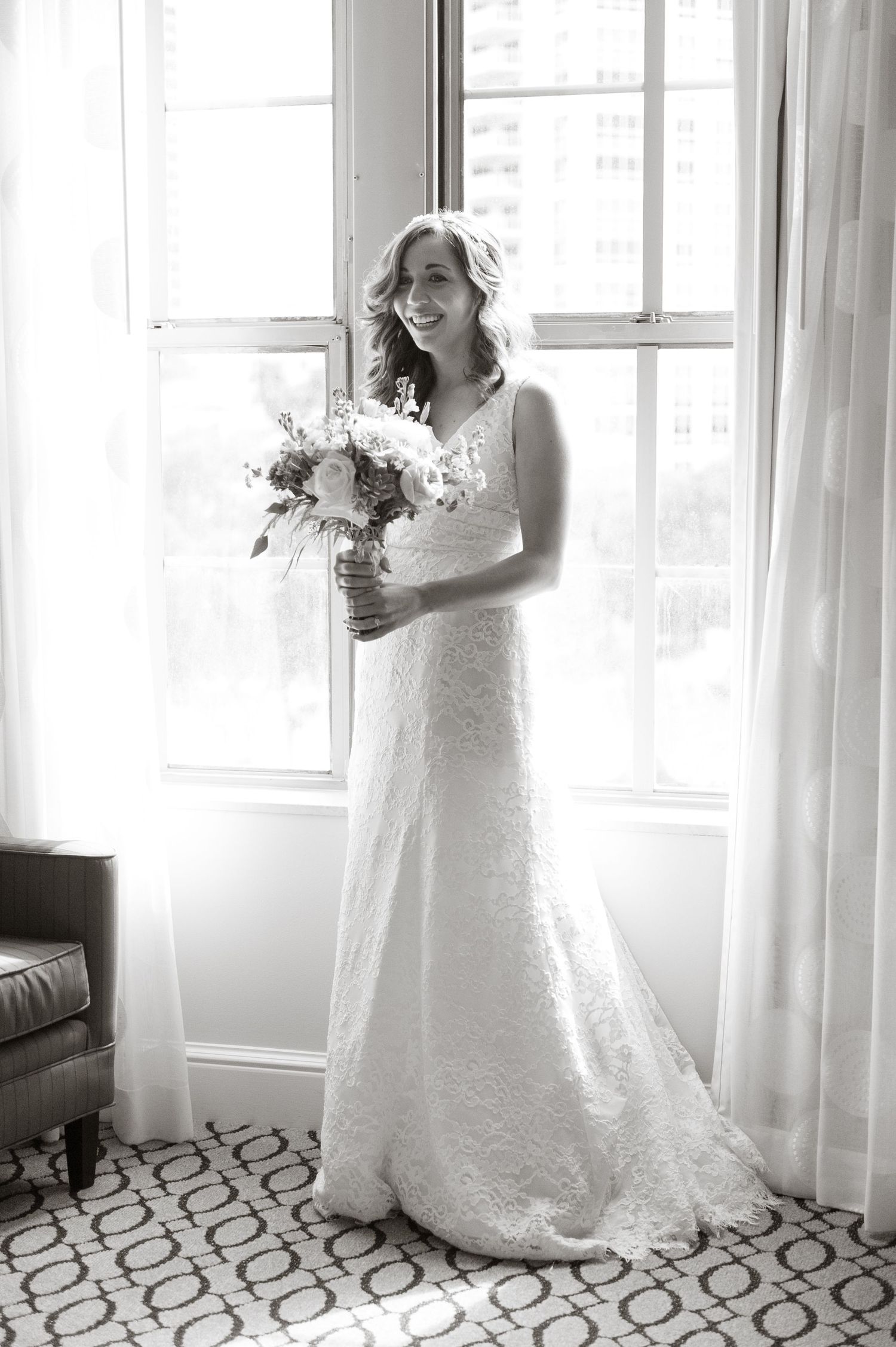 Elegant bride in a lace wedding gown smiling while holding a floral bouquet in front of a bright window.