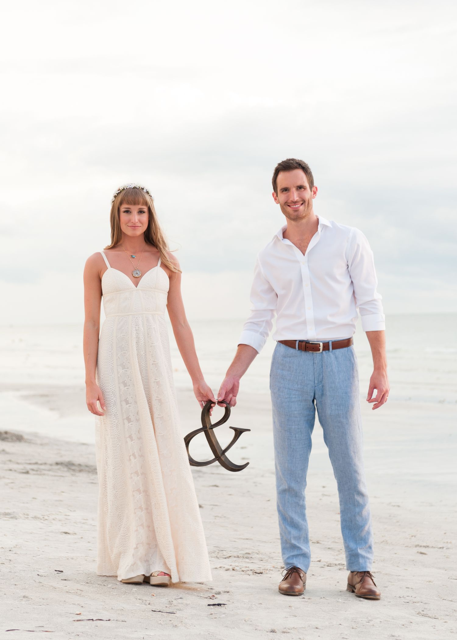 Smiling newlywed couple holds ampersand sign while standing on beach shoreline on Treasure Island, Florida.