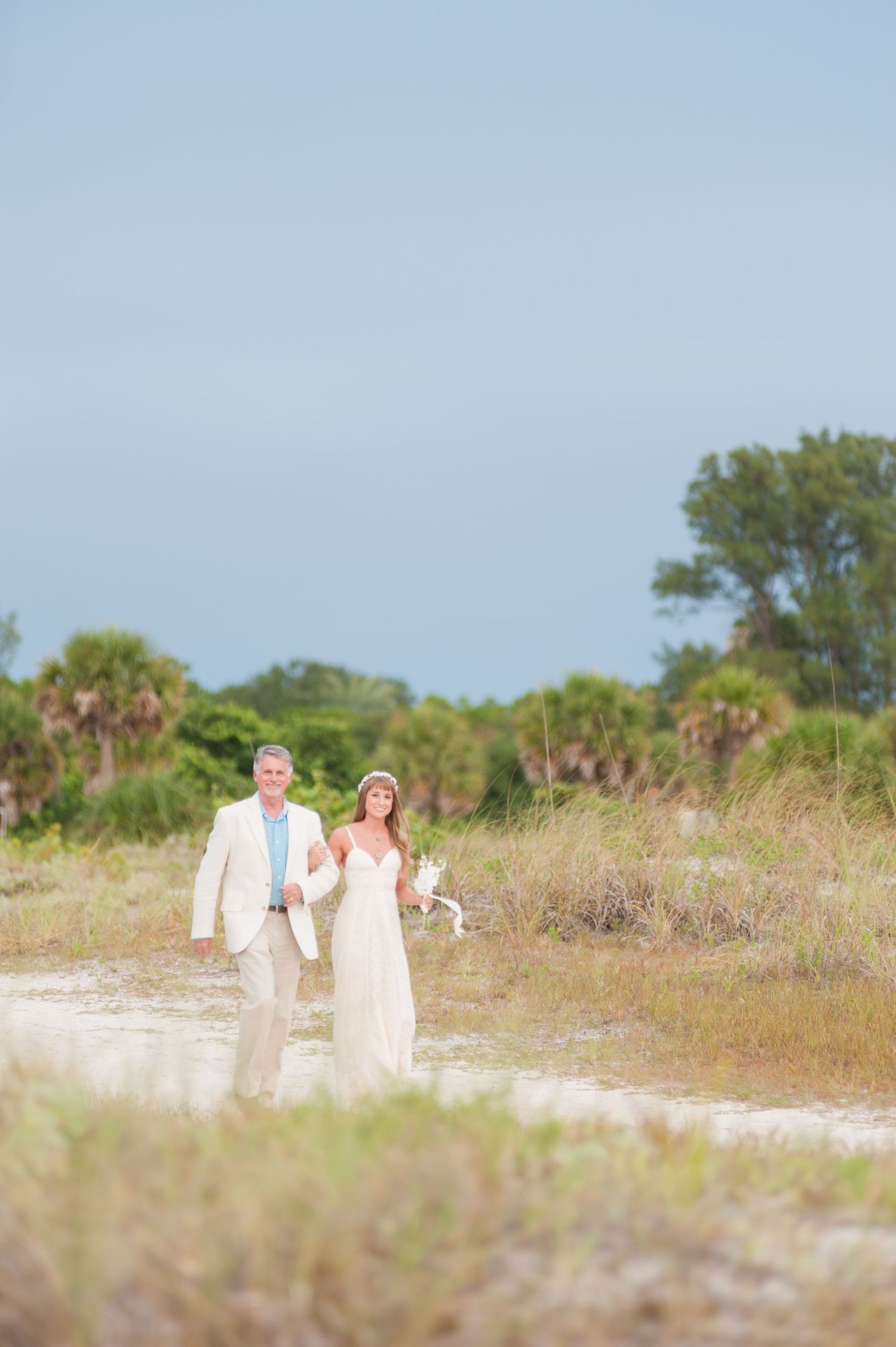 Bride and groom walk hand in hand through scenic beach landscape with palm trees behind them on Treasure Island, Florida.