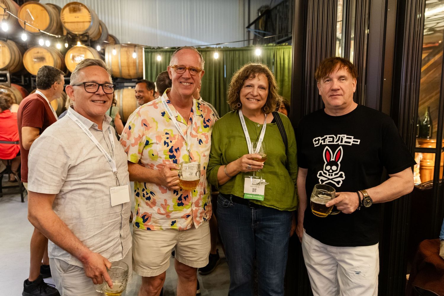 Four adults smiling and holding drinks at a brewery event with wooden barrels in the background.