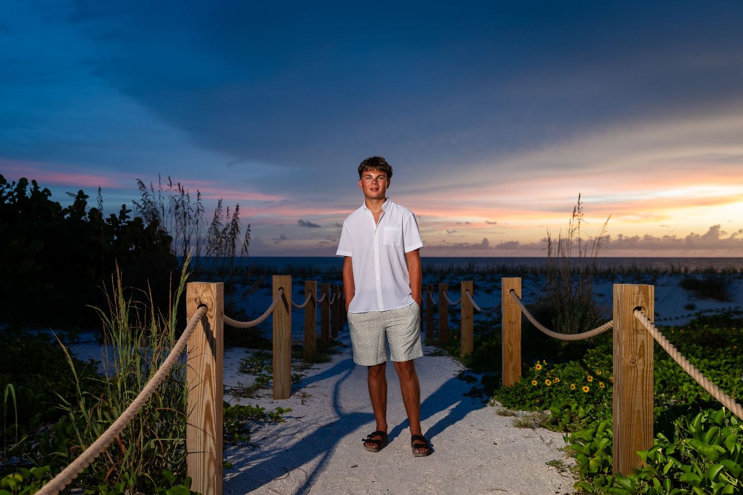 Teen boy in white shirt stands on sandy beach boardwalk at colorful sunset, rope fence lining the path.