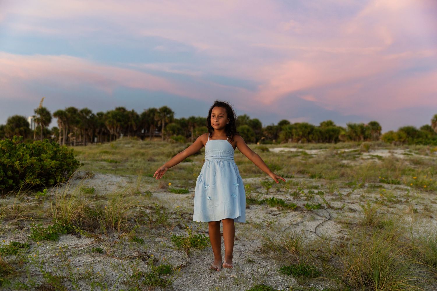A young girl in a light blue dress stands on sandy ground with palm trees and a pink sunset sky behind her.