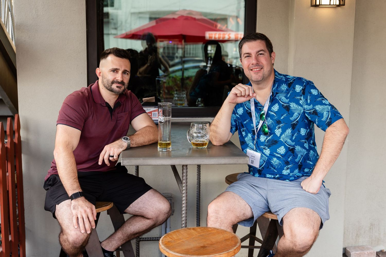 Two men smiling and sitting at an outdoor bar table with drinks, one in a blue floral shirt.