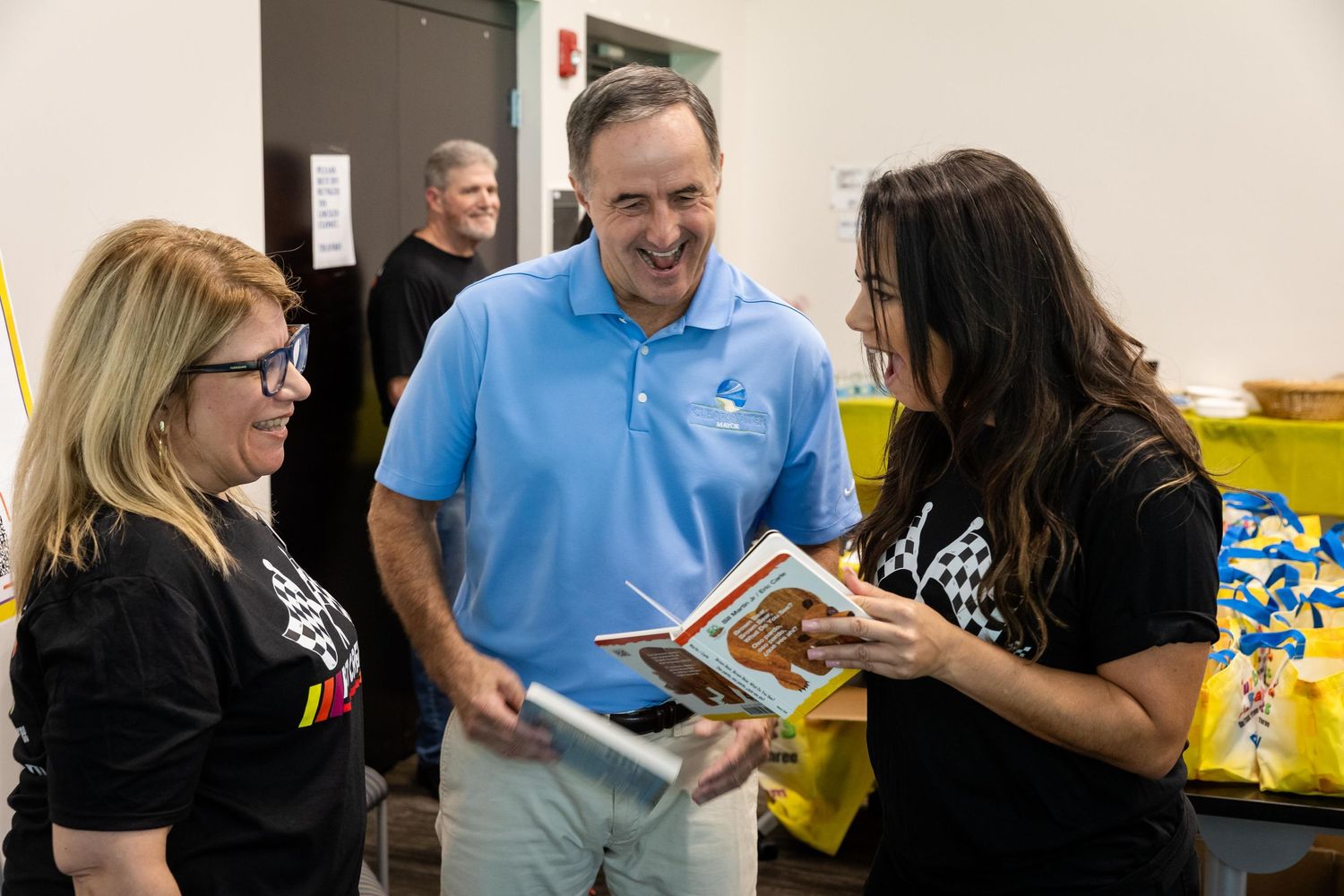 Three people smiling and looking at a book together at an indoor event, with colorful items visible in the background.