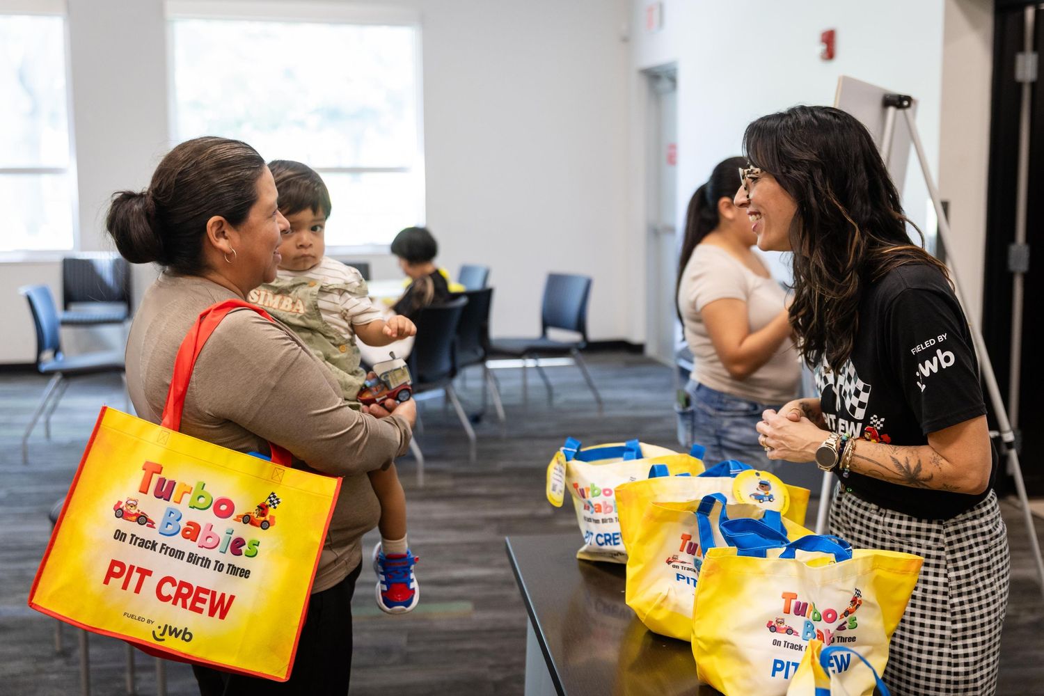 Two women chat warmly indoors, one holding a toddler, surrounded by yellow branded tote bags at a community event.