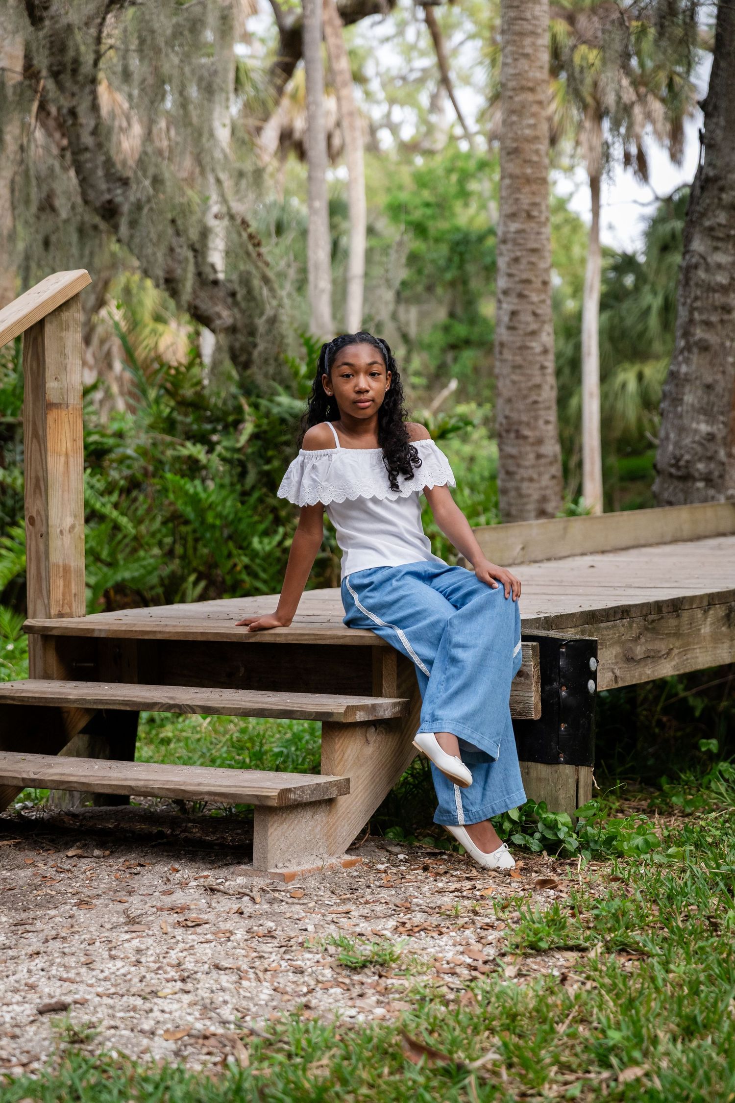 A young Black girl sits on wooden steps outdoors, wearing a white off-shoulder top and blue wide-leg pants.