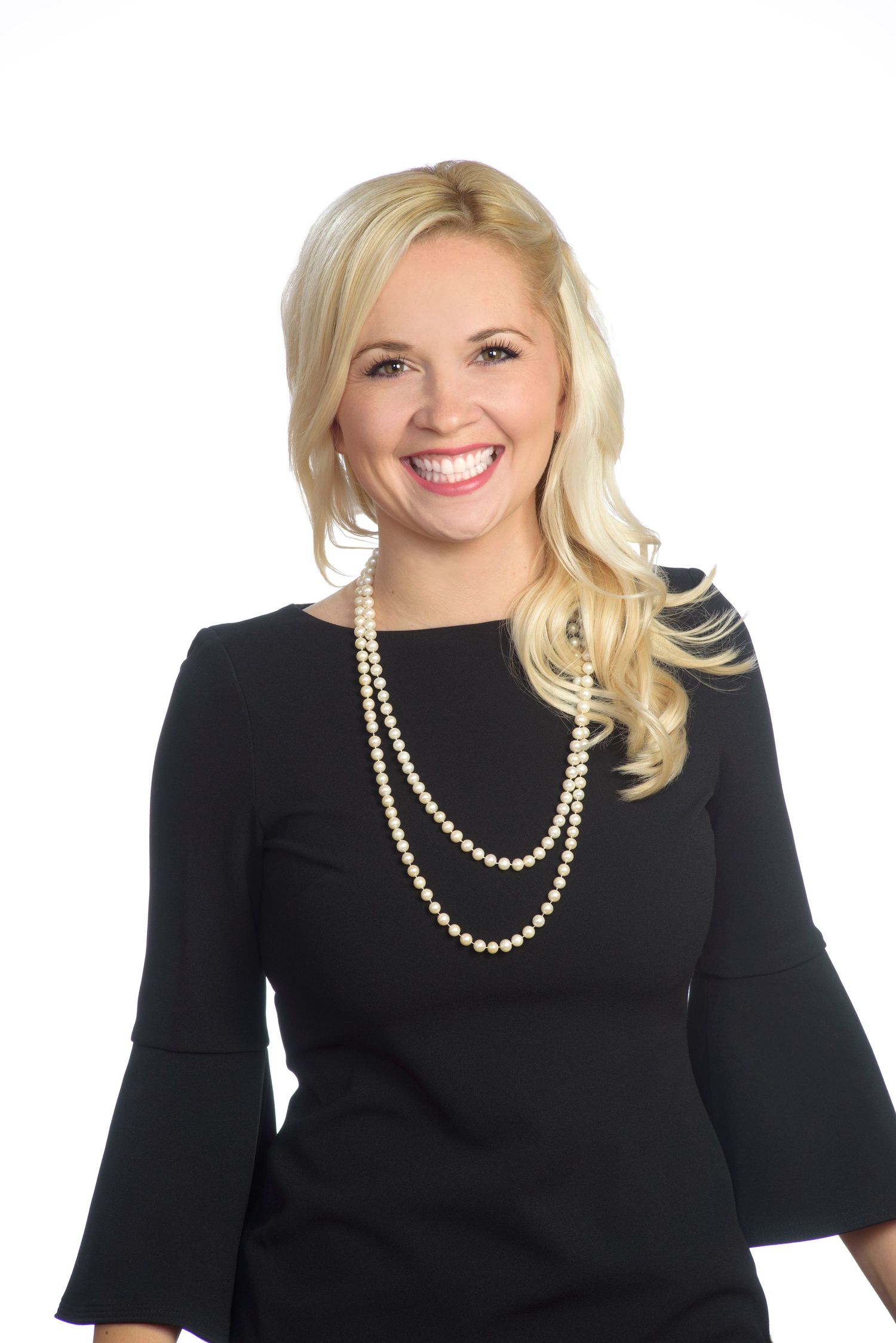 Smiling blonde woman in black dress with pearl necklace, professional headshot on white background.
